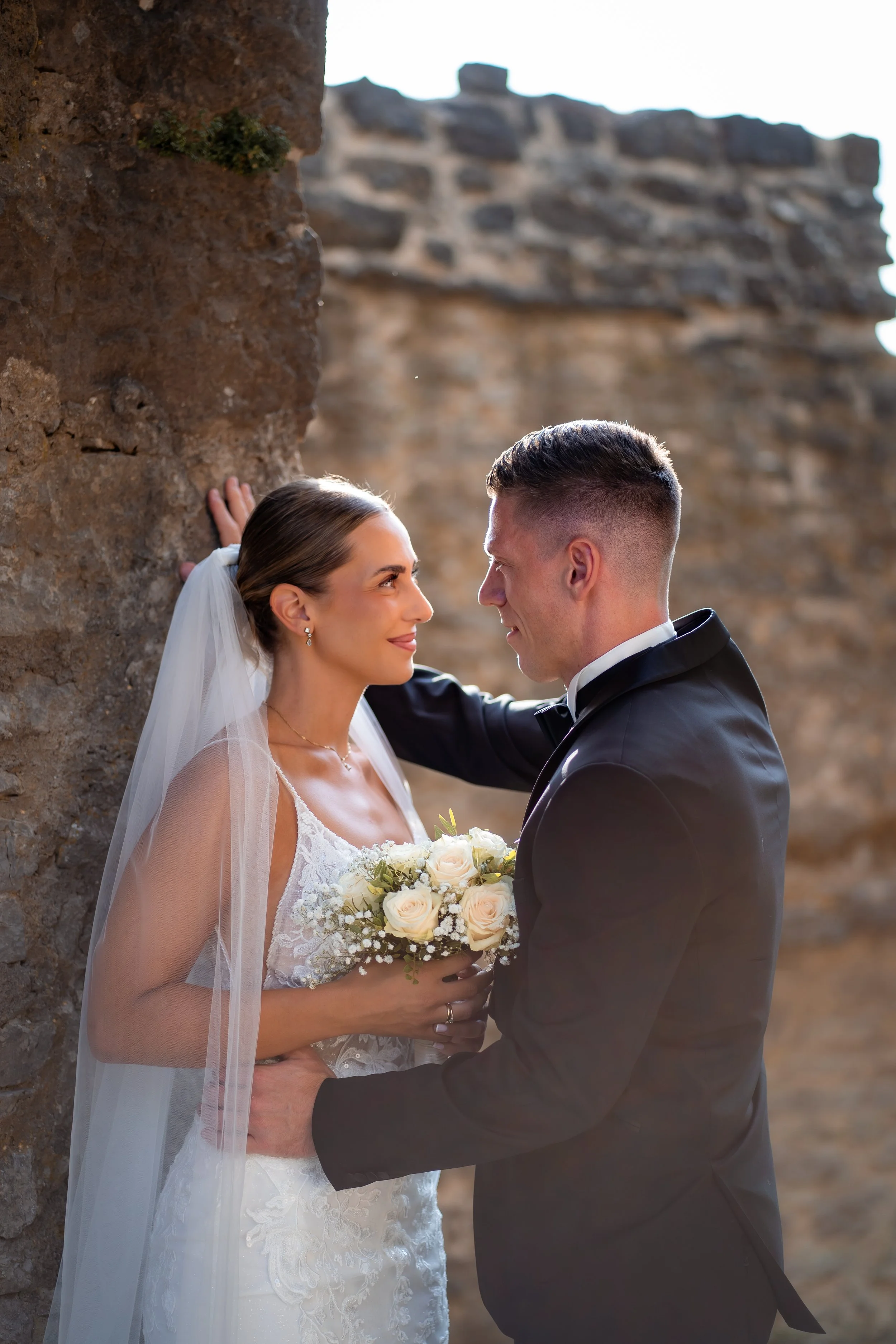 Ein Brautpaar bei einer Hochzeit, die Braut trägt ein weißes Kleid und einen Schleier, der Bräutigam trägt einen schwarzen Anzug, sie stehen sich nahe und schauen sich liebevoll an, im Hintergrund sind alte Steingebäude sichtbar.
