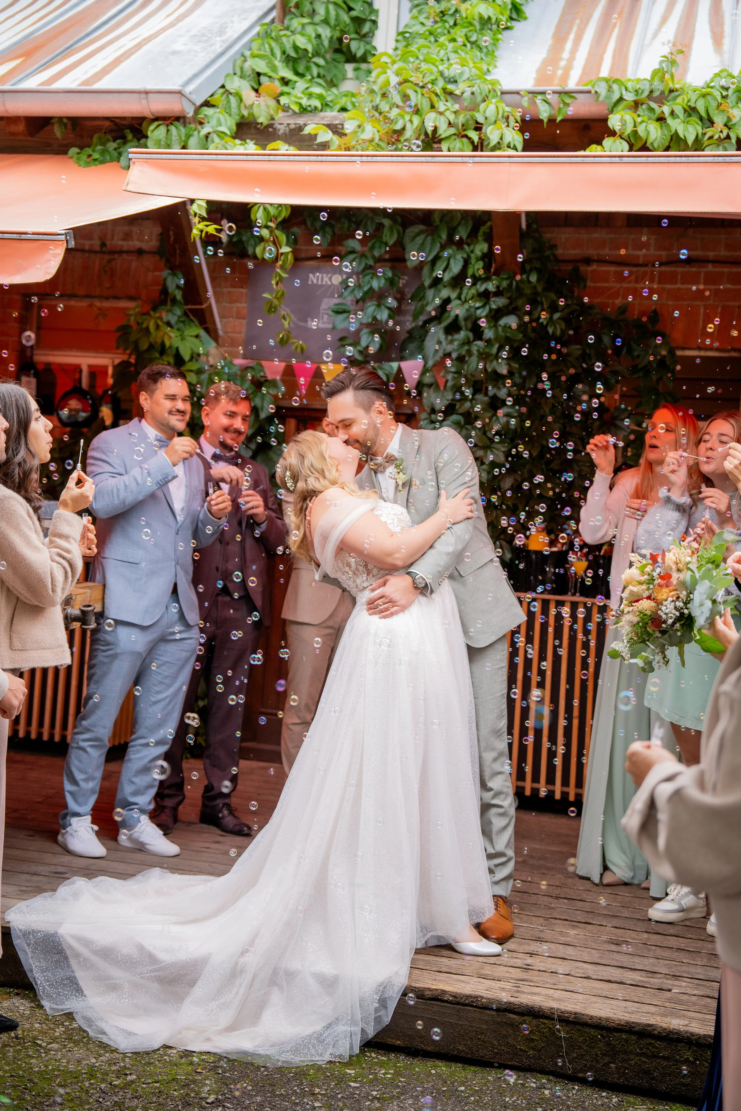 Ein Brautpaar tanzt bei einer Hochzeit, umgeben von Gästen, mit Seifenblasen im Hintergrund auf einer Holzterrasse.