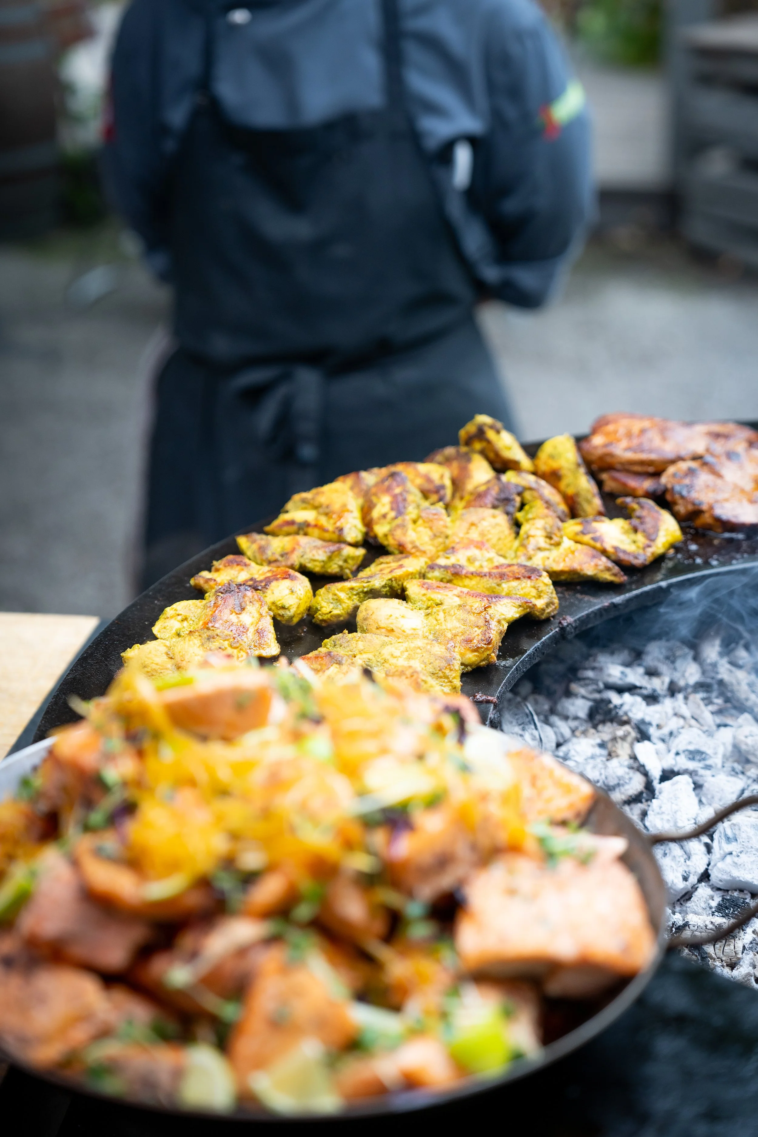 Grillplatte mit mariniertem Fleisch und Gemüse, im Hintergrund eine Person in dunkler Jacke.