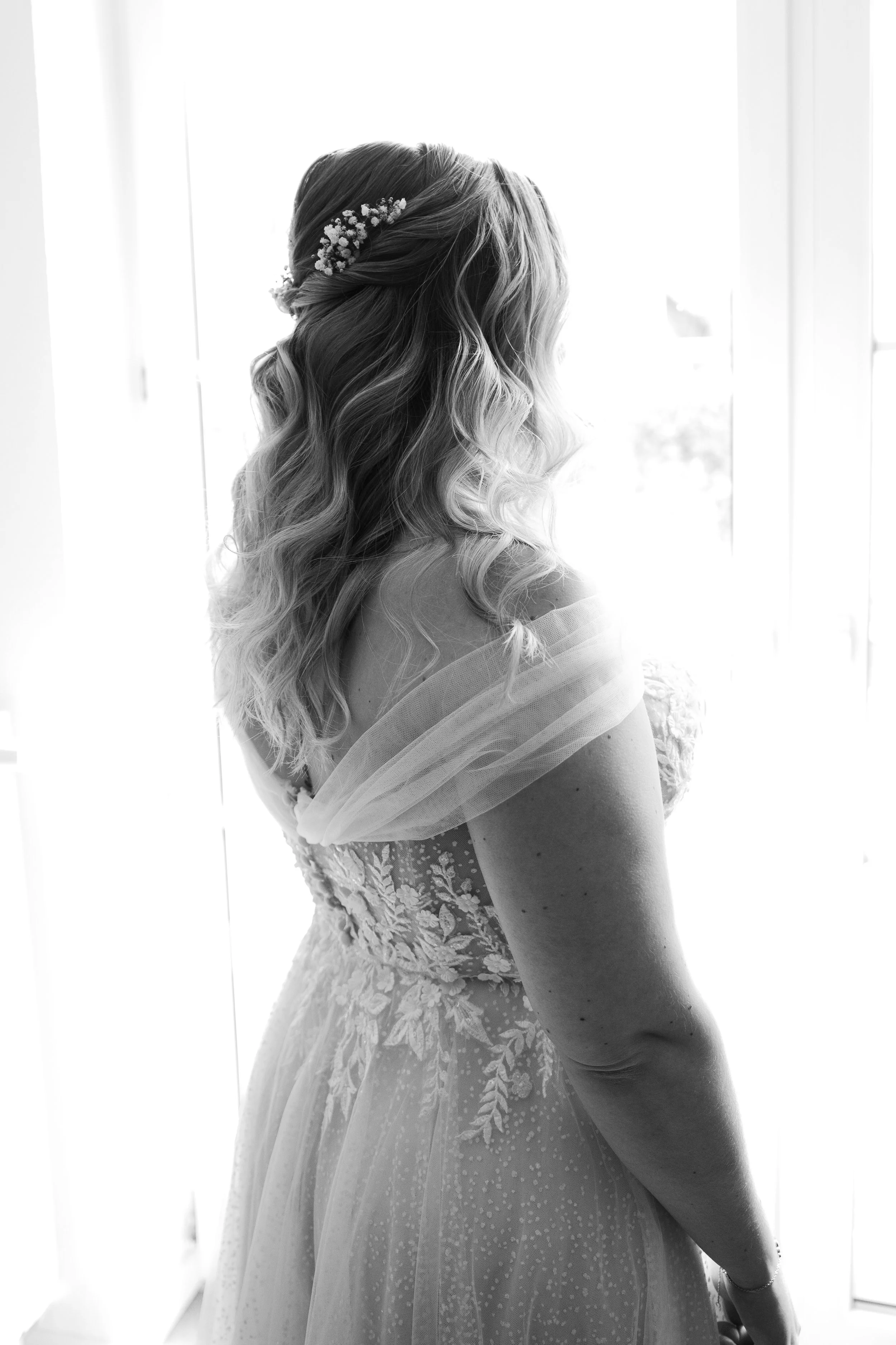 Schwarz-weiß Foto einer Frau in einem Brautkleid, die seitlich vor einem großen Fenster steht, mit geflochtenem Haar und kleinen Blumen im Haar