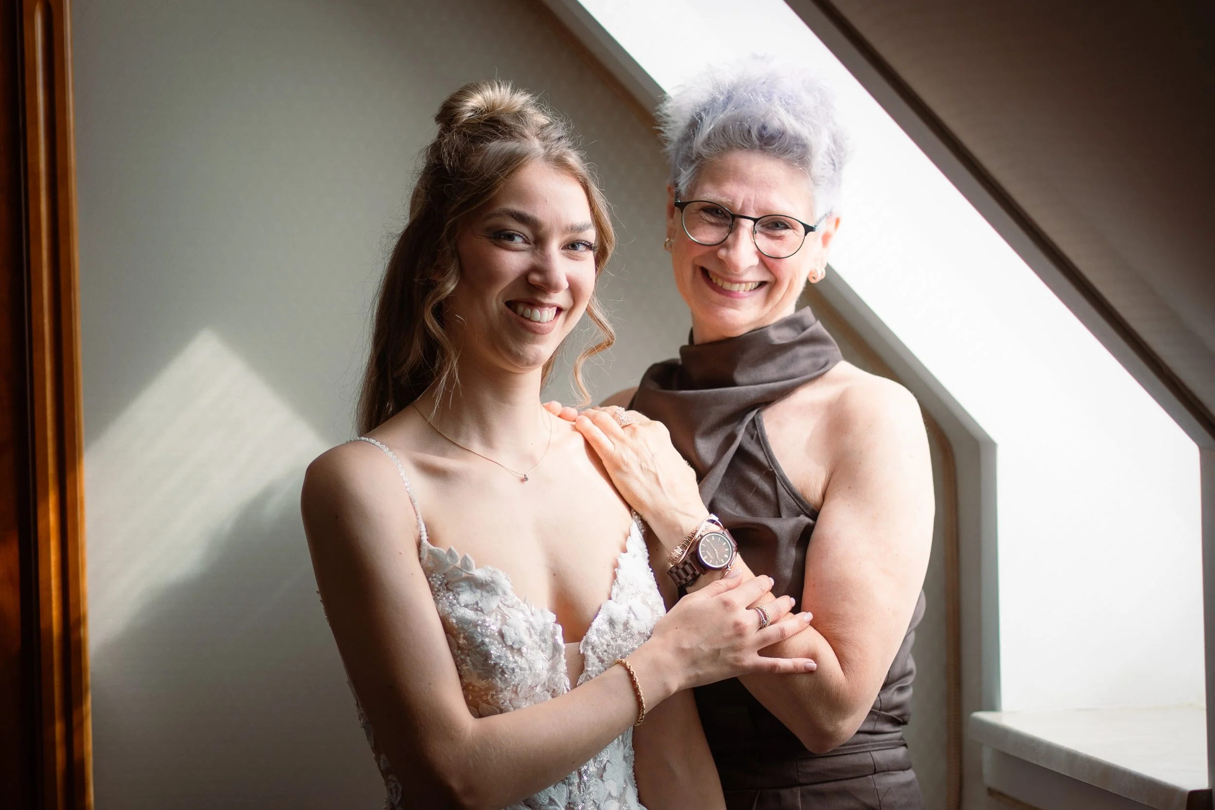 Zwei Frauen stehen nah beieinander, eine junge Frau in einem weißen Kleid und eine ältere Frau mit weißen Haaren und Brille, vor einem Fenster mit Dach schräg im Hintergrund.