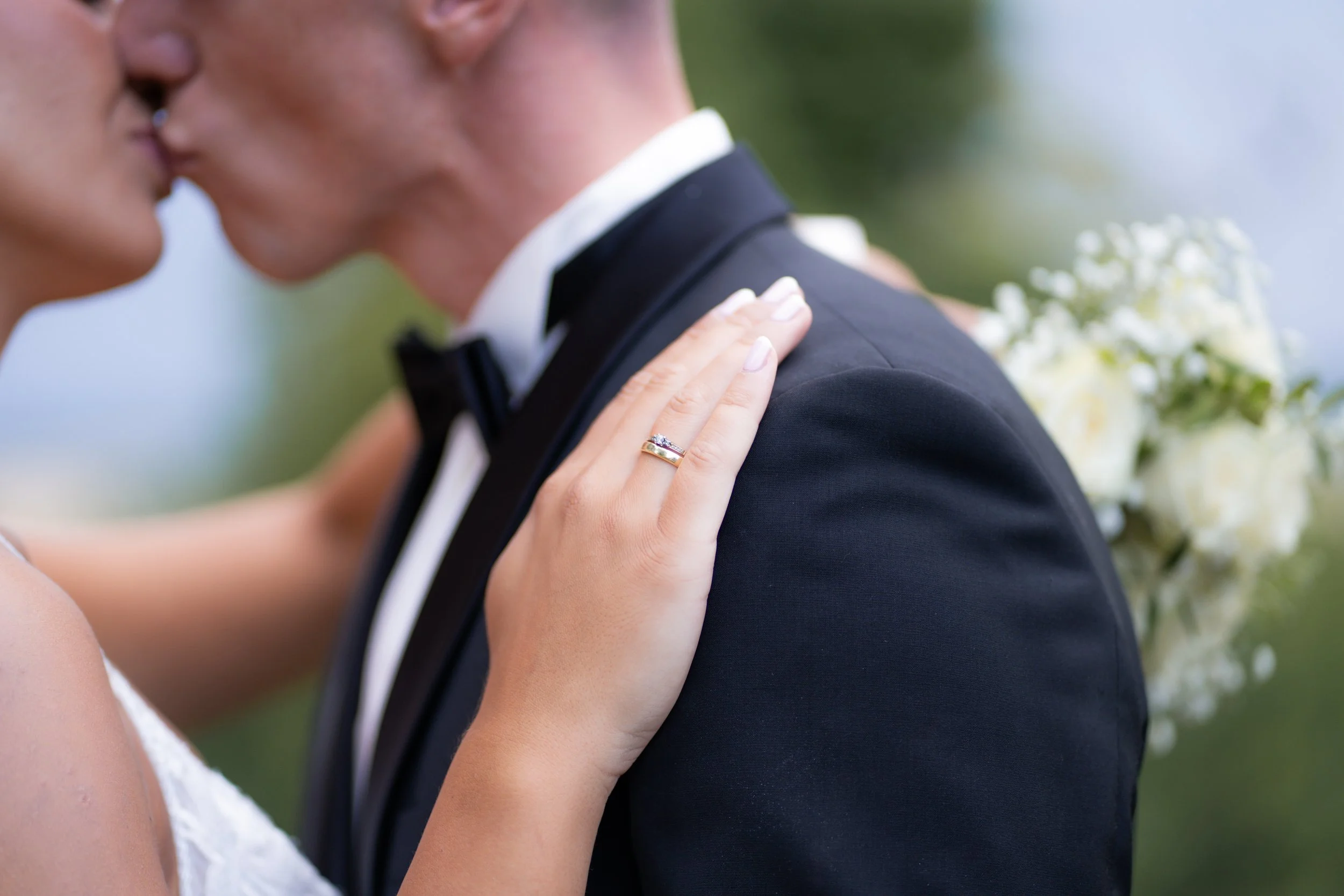 Nahaufnahme eines pärchen auf einer Hochzeit, der Blick auf die Schulter eines Mannes im schwarzen Anzug und weiße Hemd. Eine Hand eines Partners mit Ehering ruht auf seiner Schulter, eine Frau mit weißem Kleid berührt den Mann. Der Hintergrund ist u