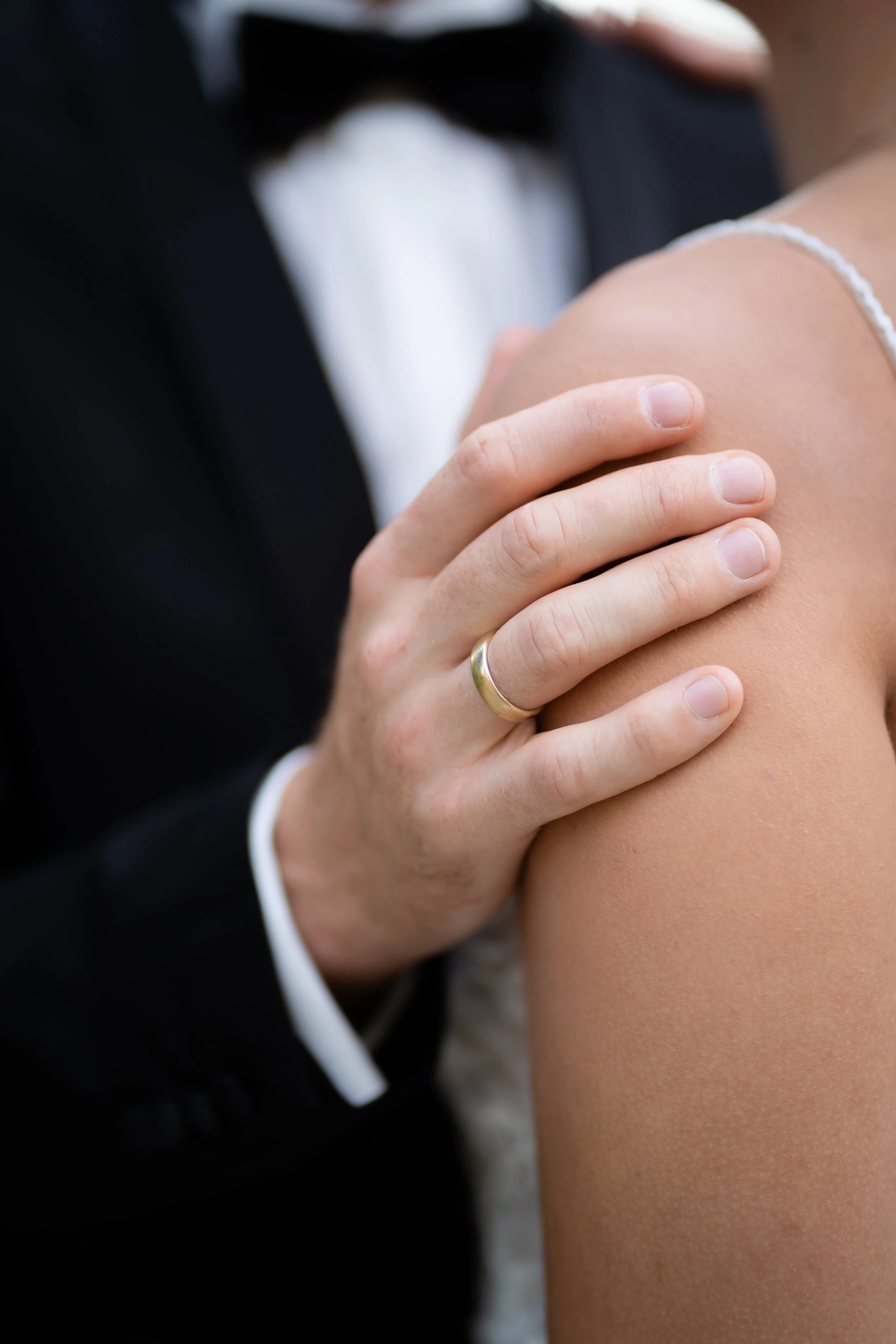 Nahaufnahme eines Mannes in einem schwarzen Anzug, der eine nackte Frau berührt, ihr Schulter mit der Hand hält, die Frau trägt eine weiße Spitze-Ärmchen. Das Bild ist verschwommen und fokussiert auf die Hand und Schulter.  Brautpaarshooting