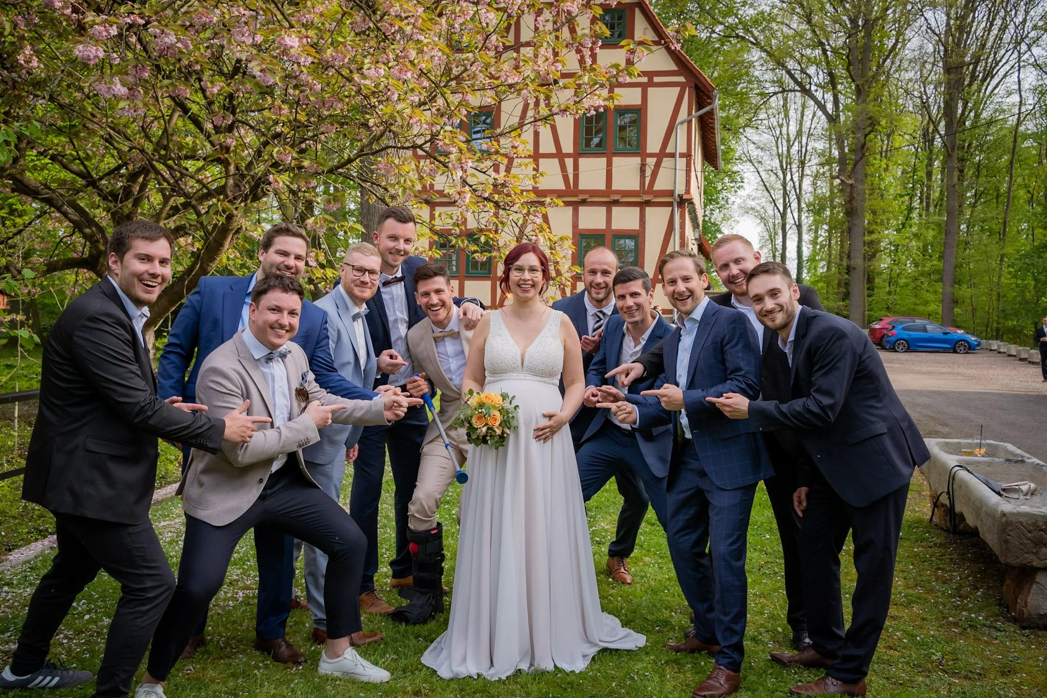 Eine Gruppe von Menschen, wahrscheinlich Freunde, bei einer Hochzeit im Garten, alle lächeln und schauen zur Kamera. Im Hintergrund sind Bäume, blühende Kirschblüten und ein Haus mit Fachwerk.