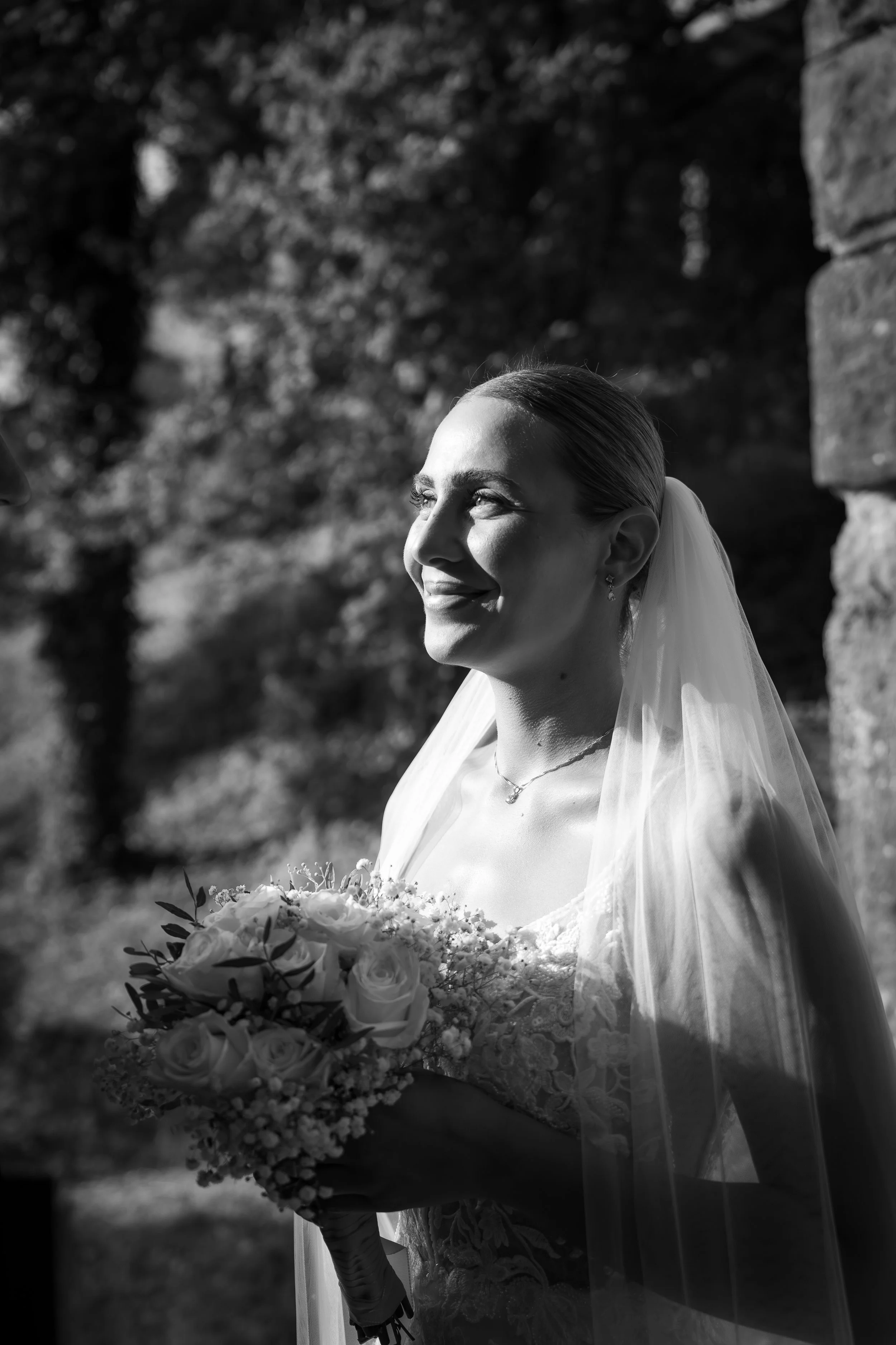 Schwarz-Weiß-Fotografie einer braut, die ein Brautkleid trägt und einen Blumenstrauß hält, mit einem Hintergund aus Bäumen. Sie lächelt und blickt zur Seite.