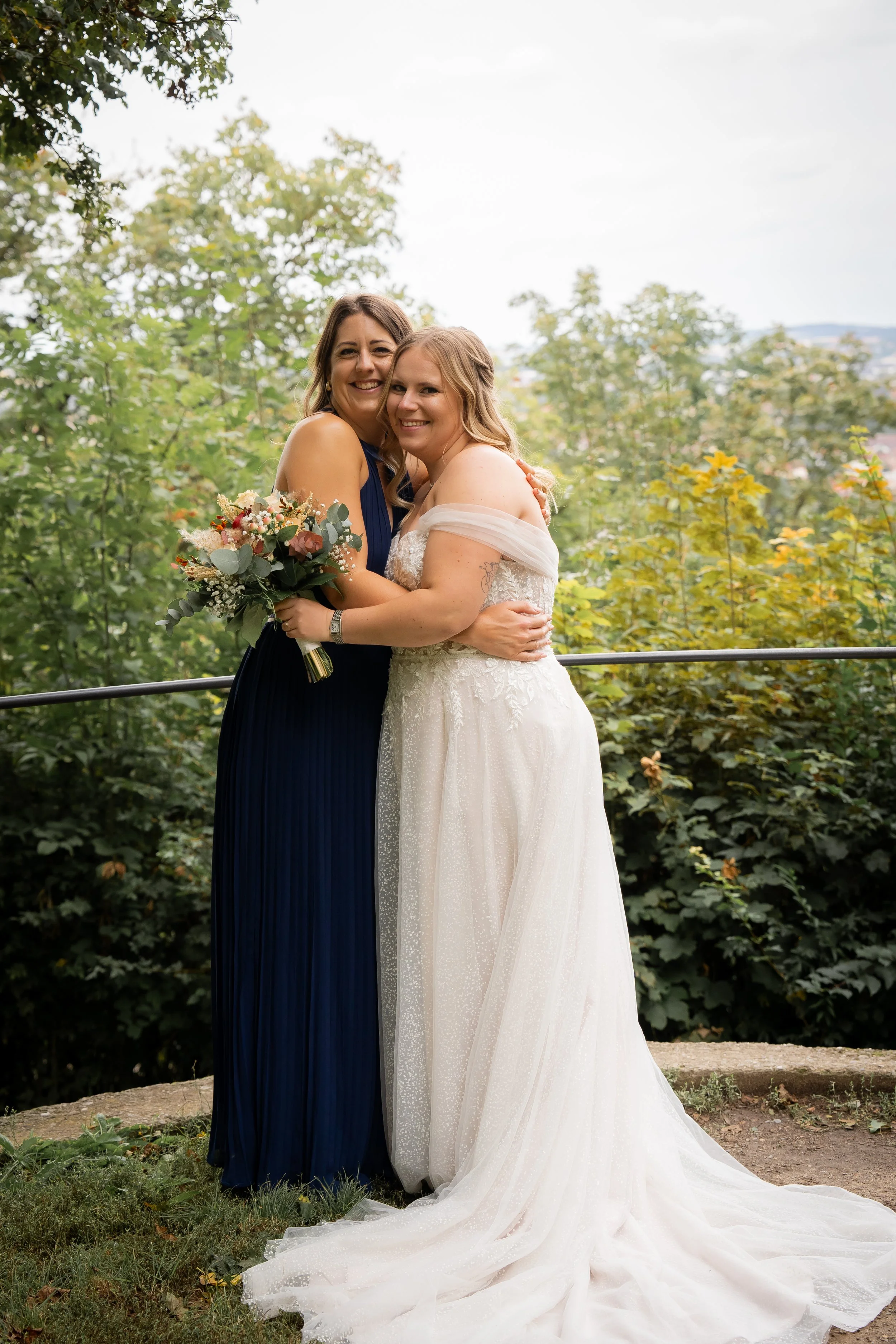 Zwei Frauen bei einer Hochzeit, umarmend vor einer grünen, bewaldeten Kulisse.