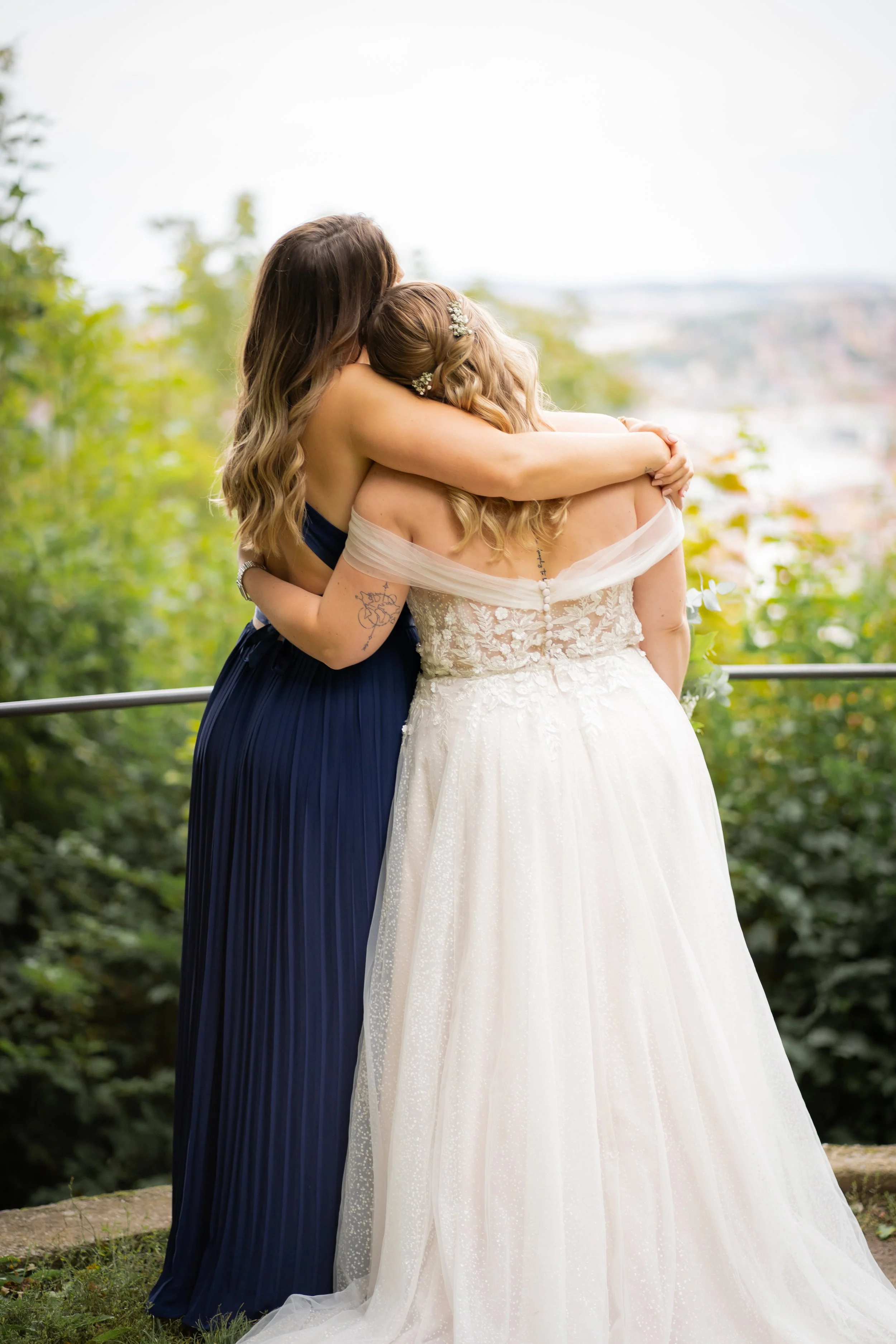 Zwei Frauen umarmen sich im Grünen bei Sonnenlicht, eine trägt ein dunkles Abendkleid und die andere ein weißes Brautkleid mit Schleier.