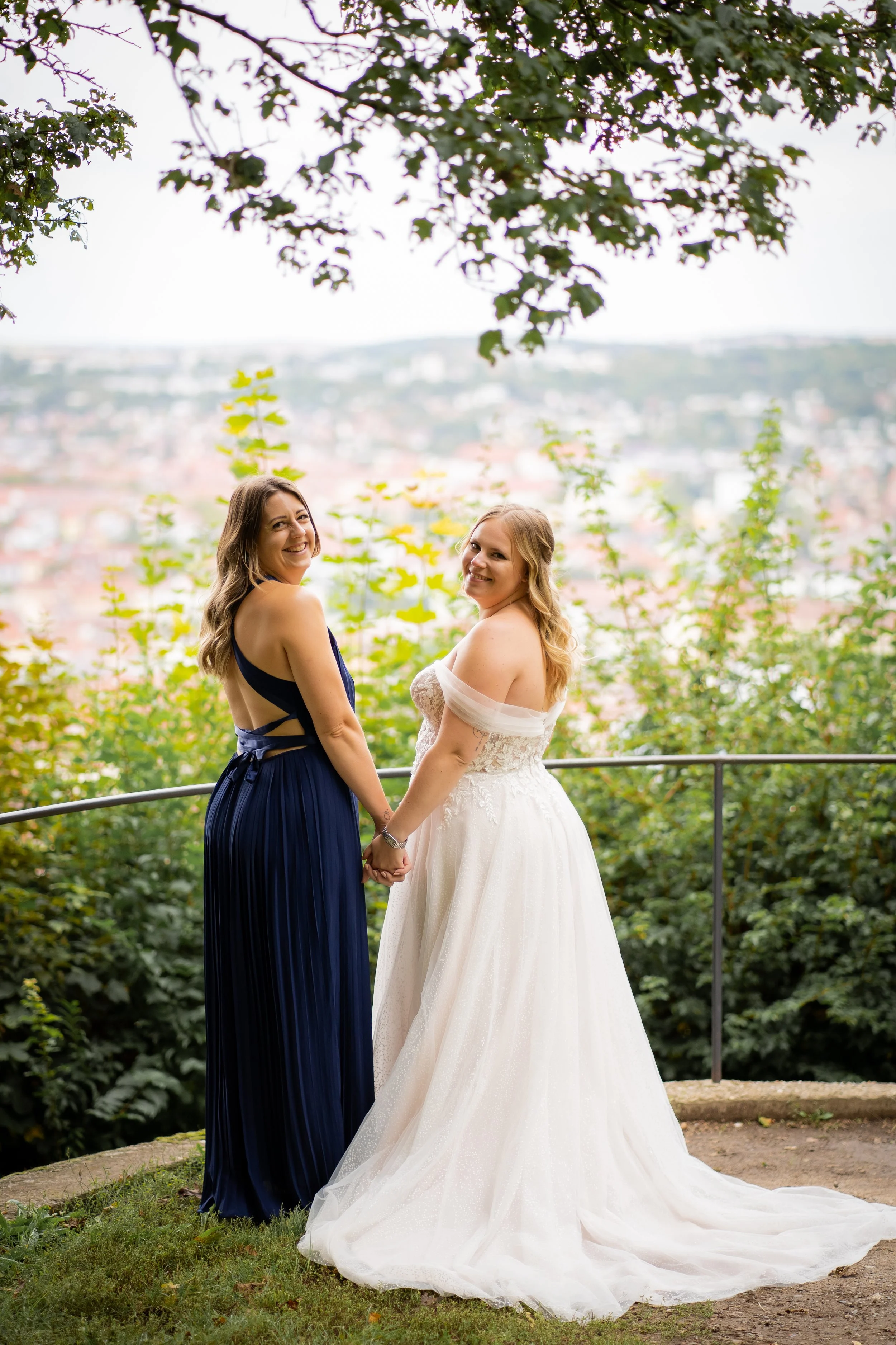 Zwei Frauen, eine in einem schwarzen Kleid und eine in einem weißen Brautkleid, halten sich an den Händen und lächeln. Sie stehen im Freien vor einer Gartenkulisse, umgeben von grünen Pflanzen und Bäumen, mit einer Stadt im Hintergrund bei Tageslicht