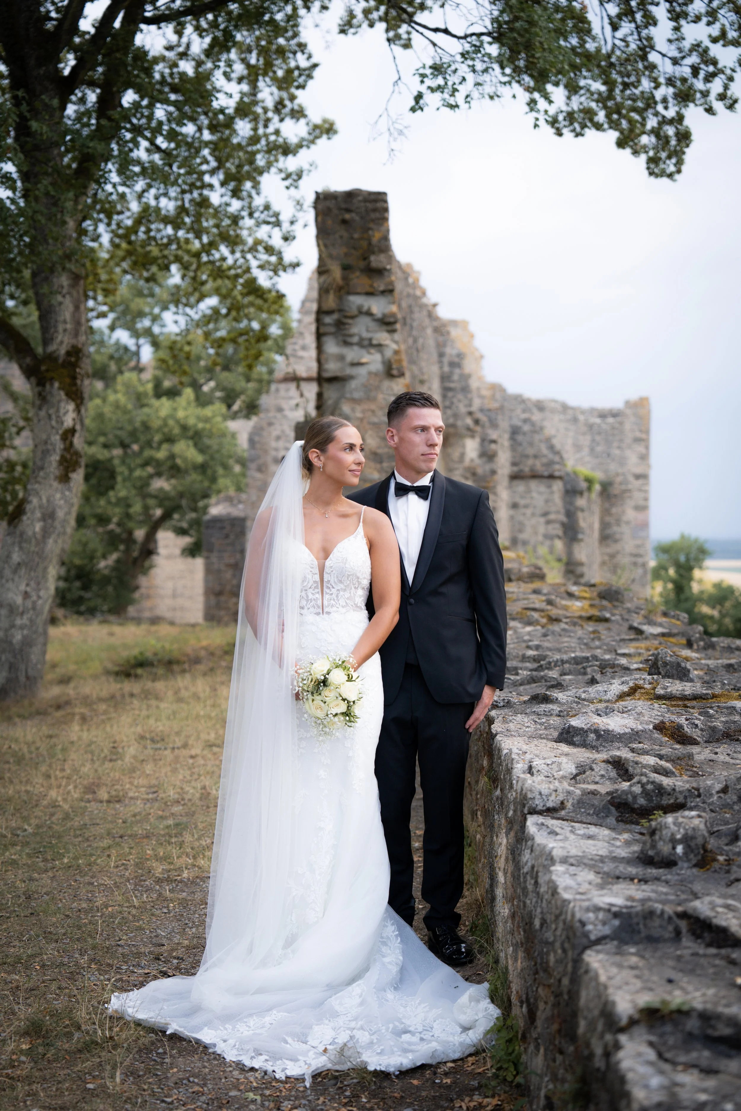 Braut und Bräutigam stehen an einer alten Stemauer vor den Überresten einer Burg, umgeben von Bäumen, bei Sonnenuntergang, in formeller Hochzeit Kleidung.