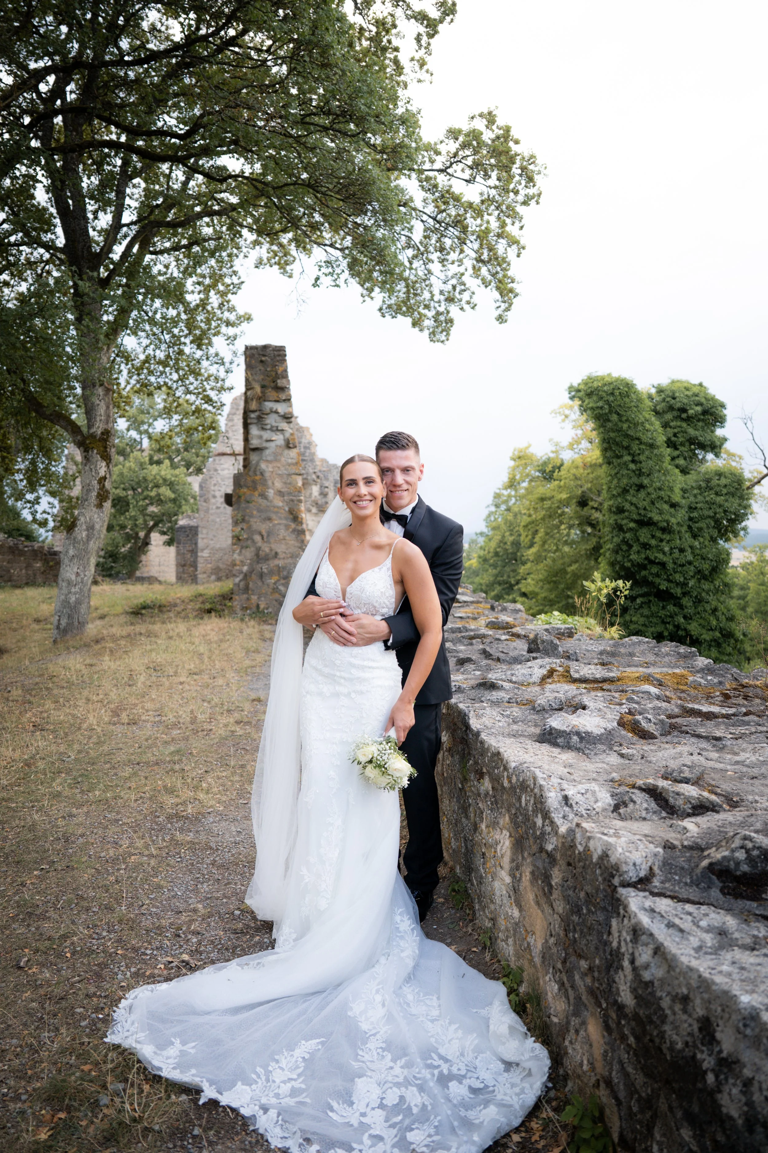 Ein glückliches Brautpaar posiert im Freien vor einer alten Steinmauer, umgeben von grünen Bäumen und historischen Ruinen, bei einer Hochzeit.