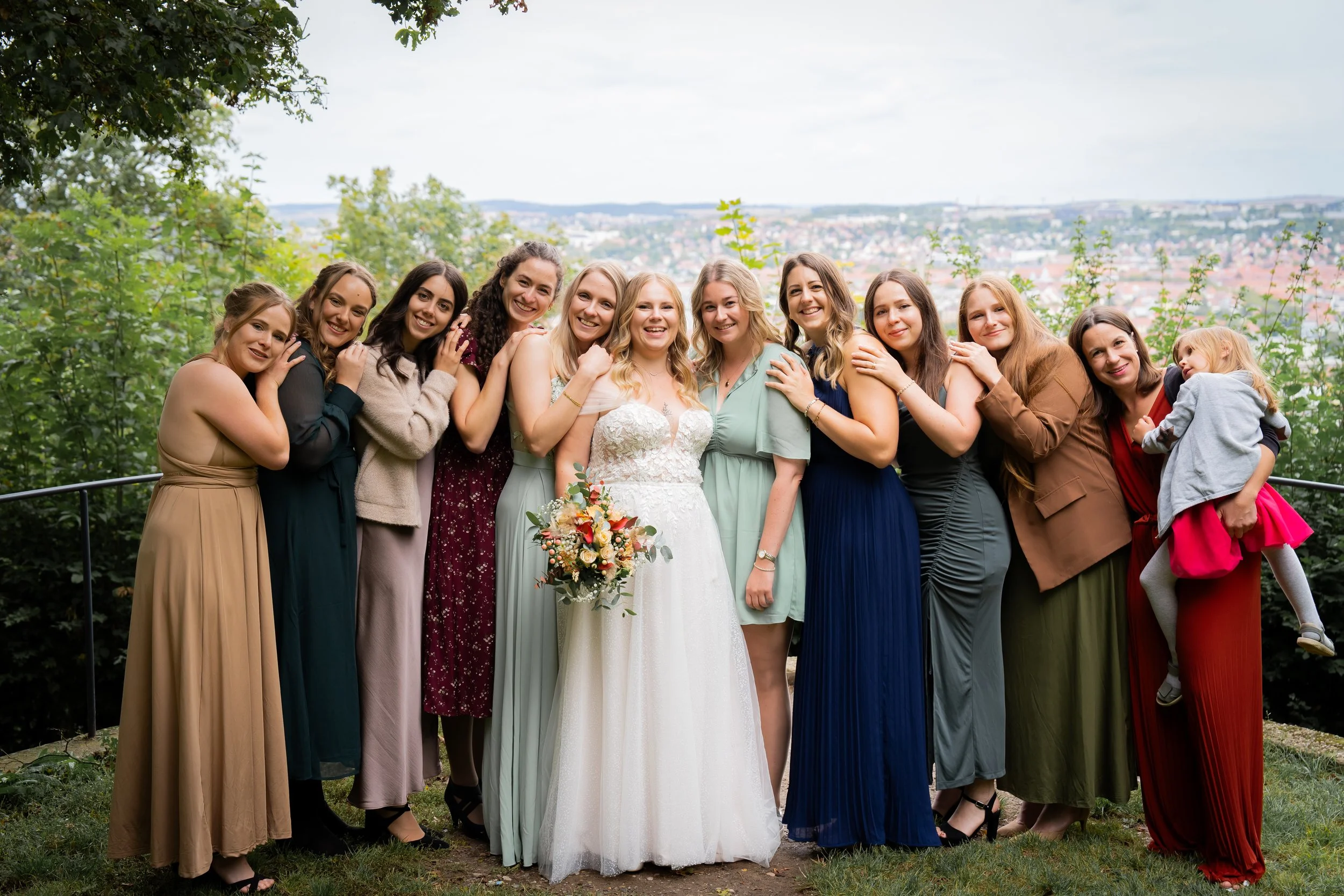 Eine Gruppe von Frauen, darunter eine Braut in einem weißen Kleid mit Brautstrauß, steht eng beieinander im Freien vor einer Landschaft mit Bäumen und Stadt im Hintergrund.