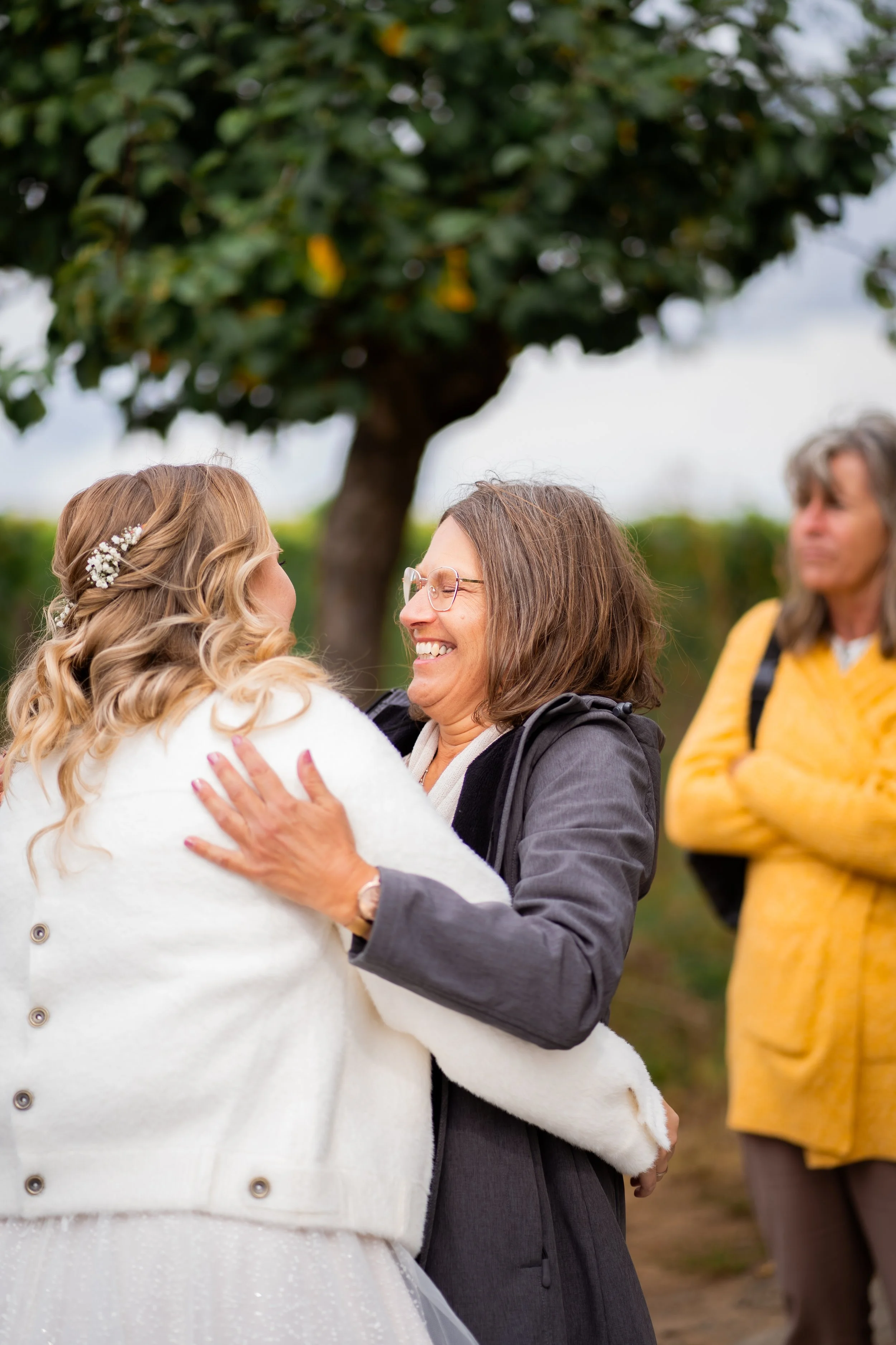 Zwei Frauen, die sich umarmen und lächeln, draußen vor einem Baum, während eine Beobachterin im Hintergrund steht.