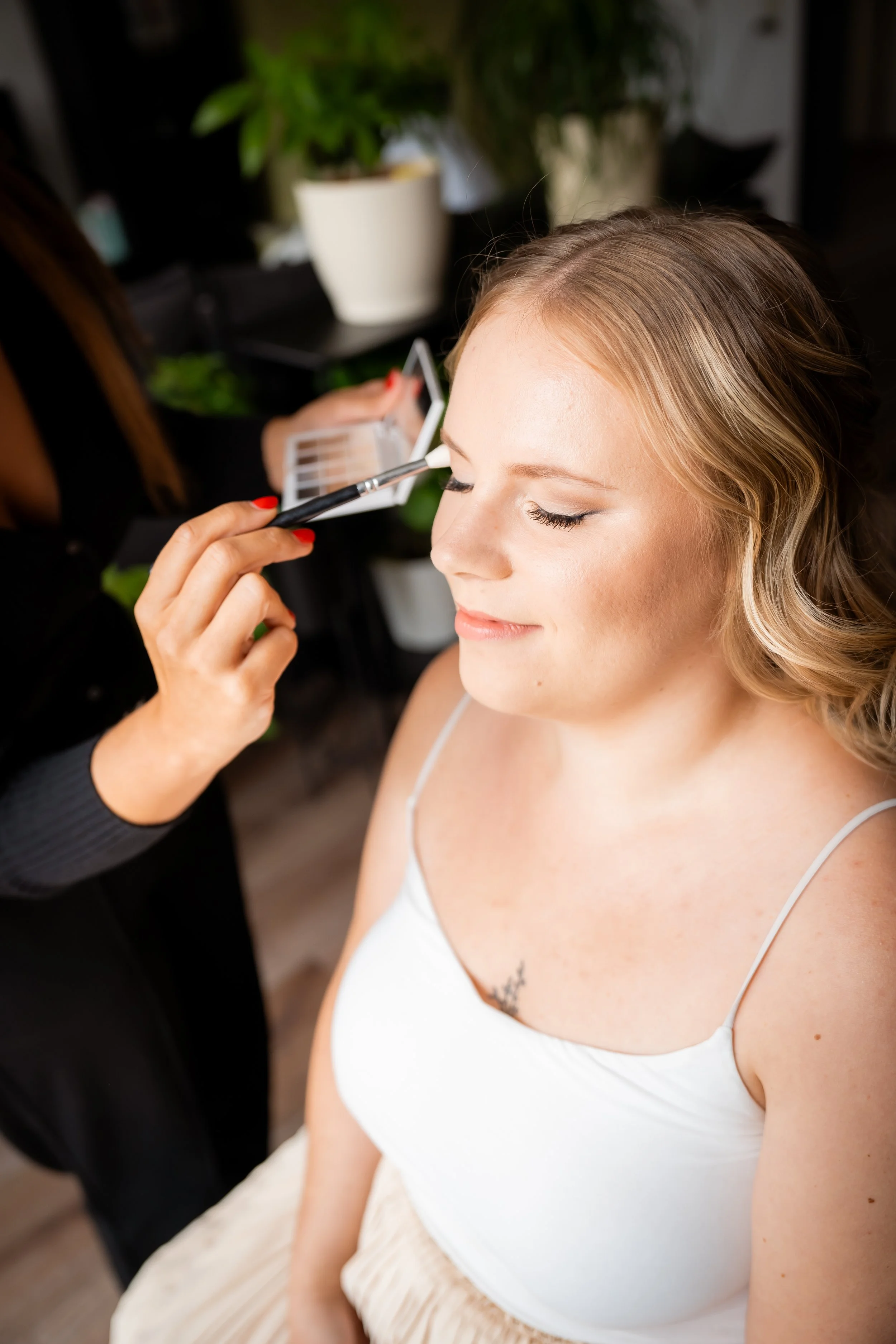 Junge Frau bekommt Make-up aufgetragen, geschlossene Augen, helle Kleidung, im Hintergrund Pflanzen.