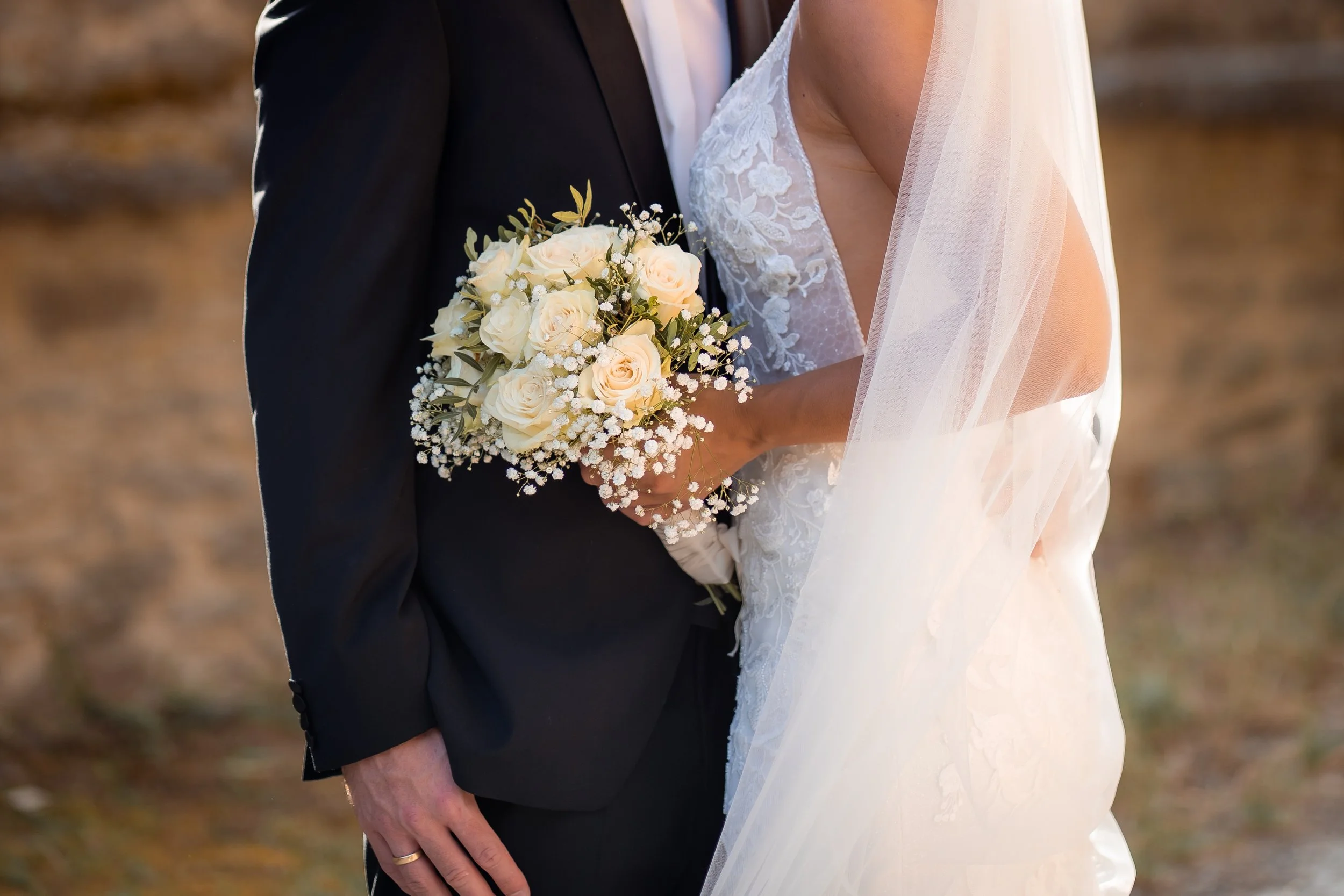 Nahaufnahme eines Brautpaares bei der Hochzeit, mit Fokus auf die Braut in einem weißen Kleid mit Spitzen und den Bräutigam im schwarzen Anzug, der eine weiße Bluse trägt. Die Braut hält einen Blumenstrauß aus weißen Rosen und kleinen weißen Blüten. Das Bild zeigt nur die obere Körperhälfte der Paare.
