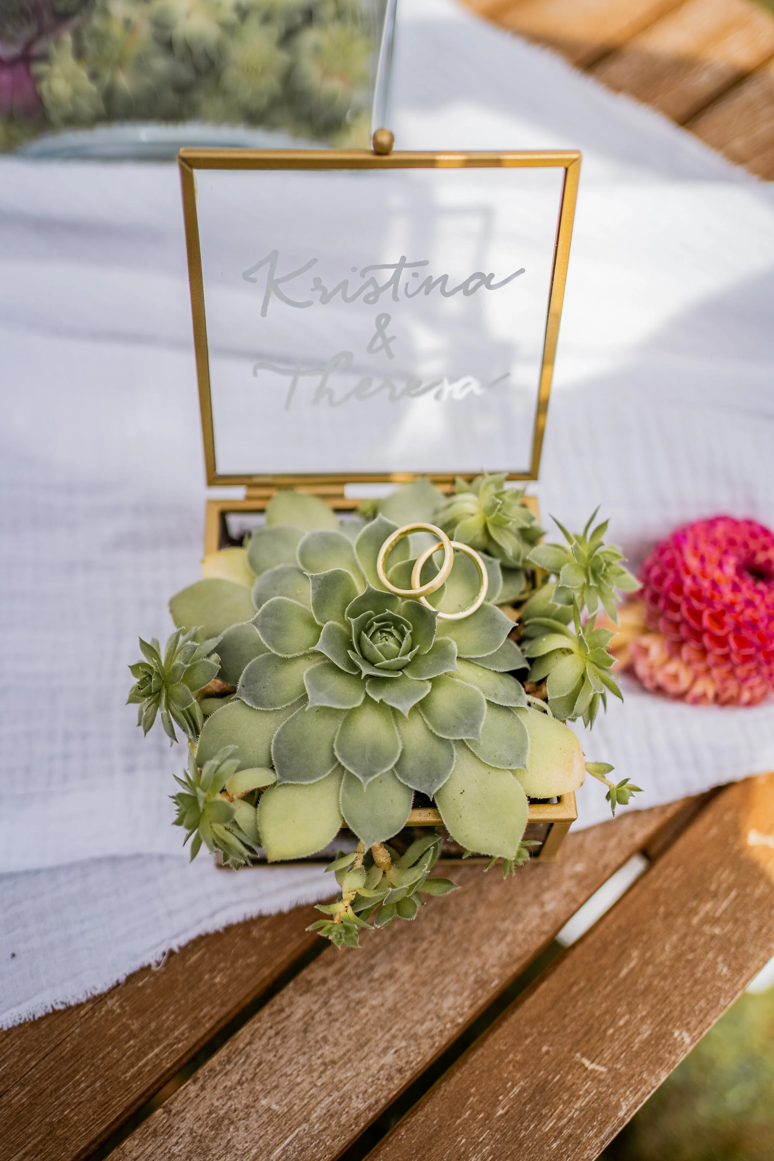 Ein dekorativer Rahmen mit den Namen 'Kristina & Theresia' auf einem weißen Tuch, umgeben von Sukkulenten und Blumen auf einem Holztisch.