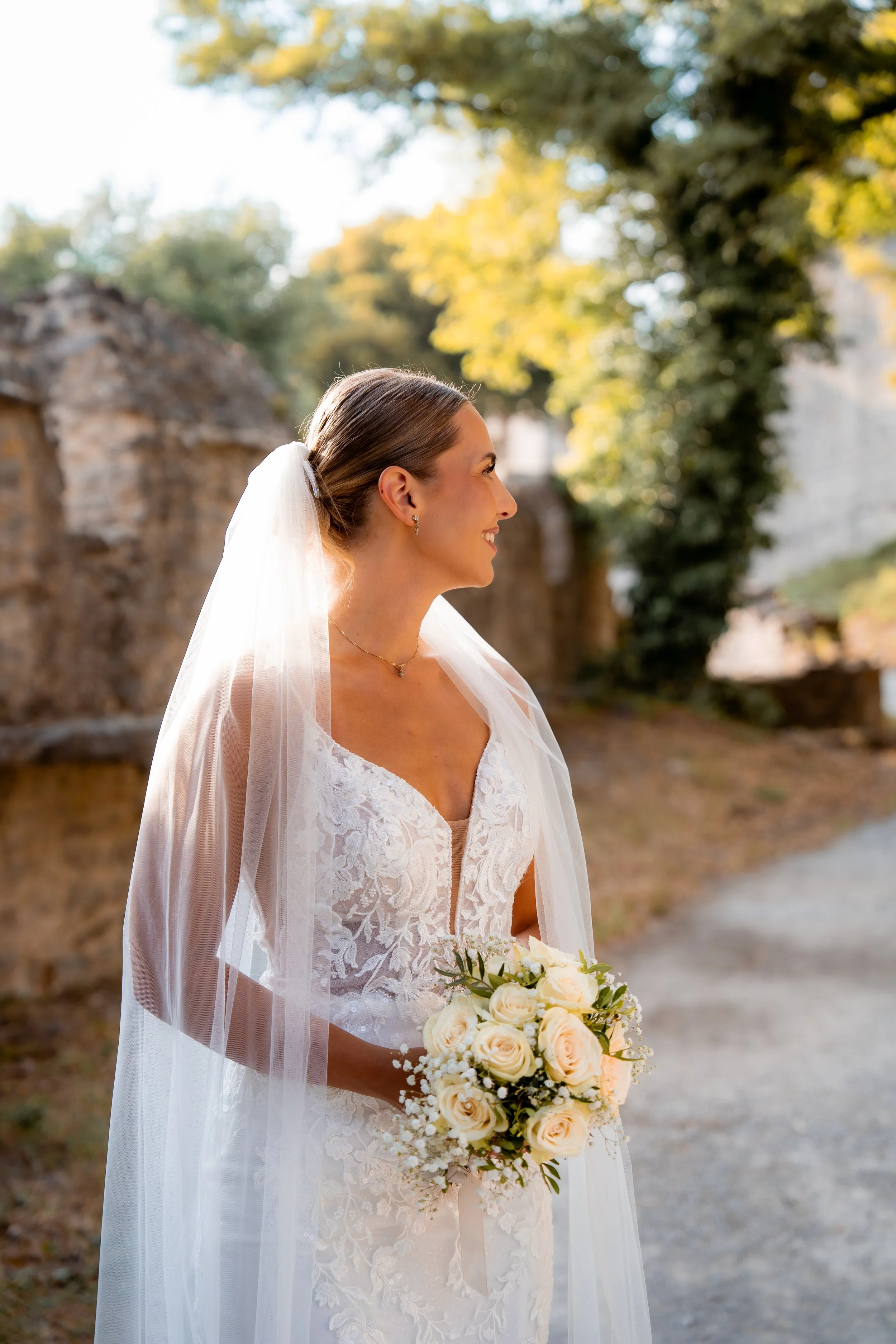 Braut mit Brautstrauß im Freien, lächelnd, bei Sonnenuntergang, trägt ein weißes Spitzenkleid und einen Schleier, im Hintergrund Bäume und eine alte Steinmauer.