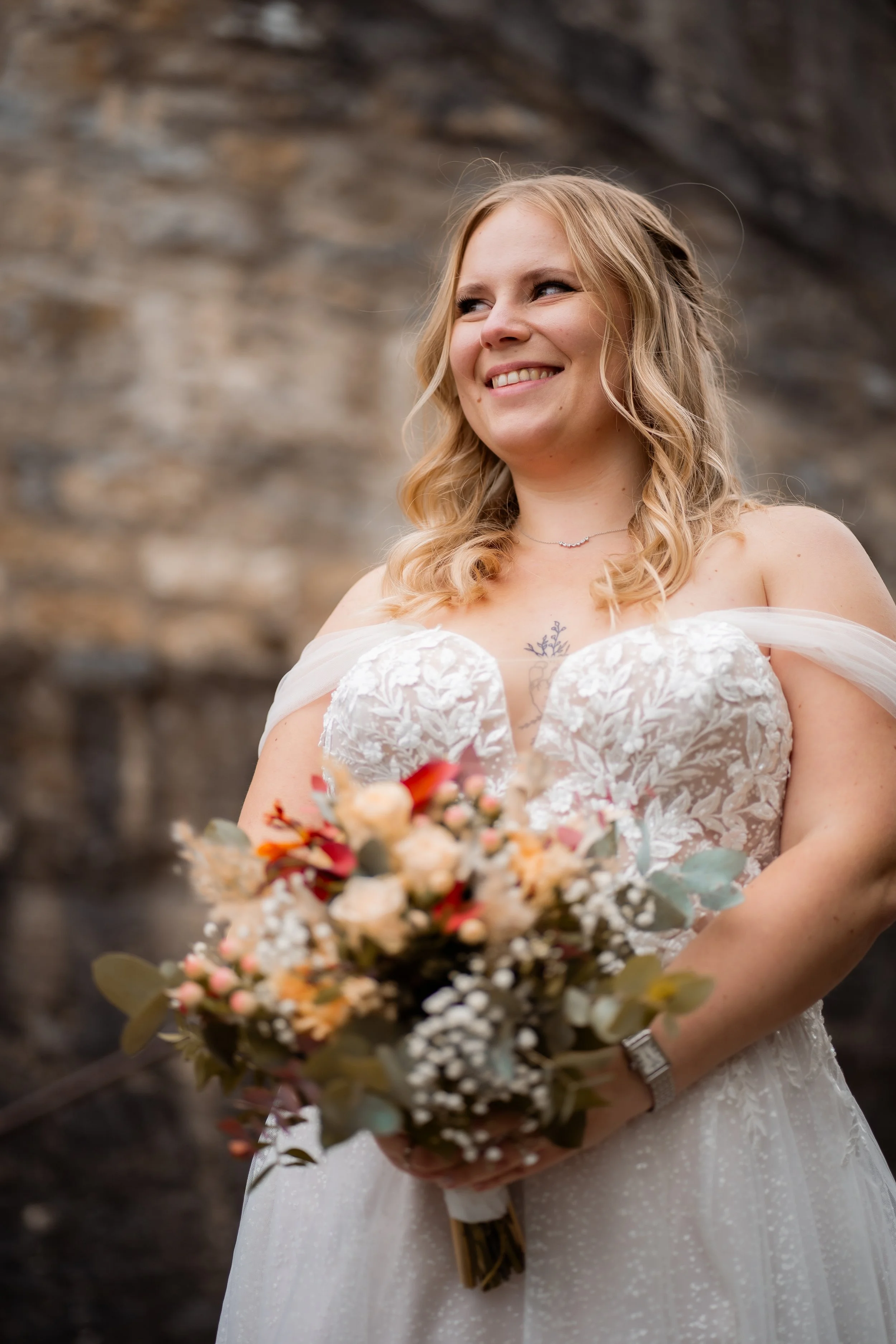 Eine lachende Frau in einem weißen Brautkleid hält einen Blumenstrauß mit verschiedenen Blumen und Eukalyptus. Der Hintergrund ist eine Steinmauer.