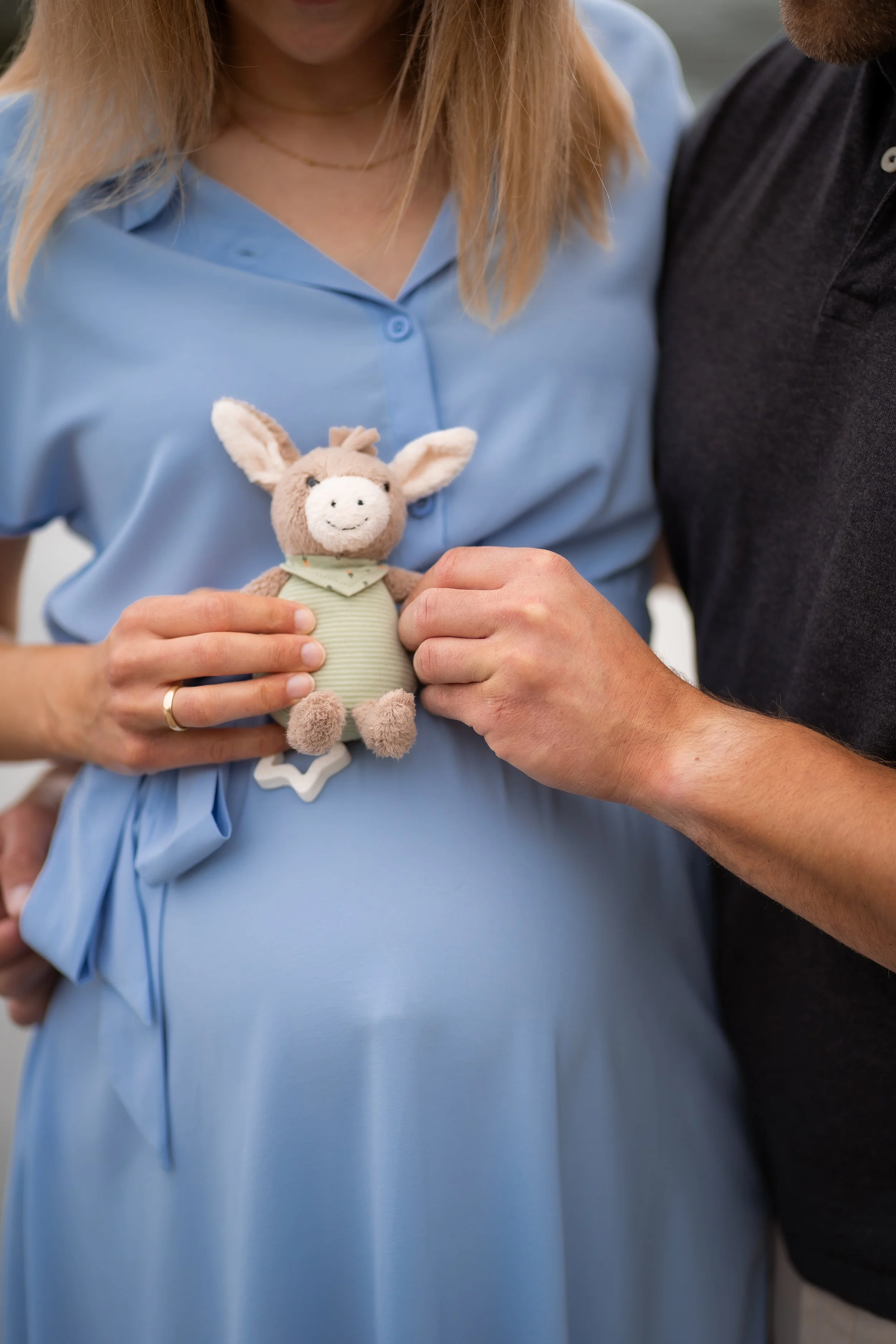 Ein Paar hält ein Plüschtier in Form eines niedlichen Eselfreunds, während sie sich nahe kommen, symbolisch für eine enge Beziehung oder ein Baby. Die Frau trägt ein hellblaues Kleid und eine goldene Halskette. Die Details im Hintergrund sind unscharf, Fokus liegt auf den Händen, dem Plüschtier und den Oberkörpern der Personen.