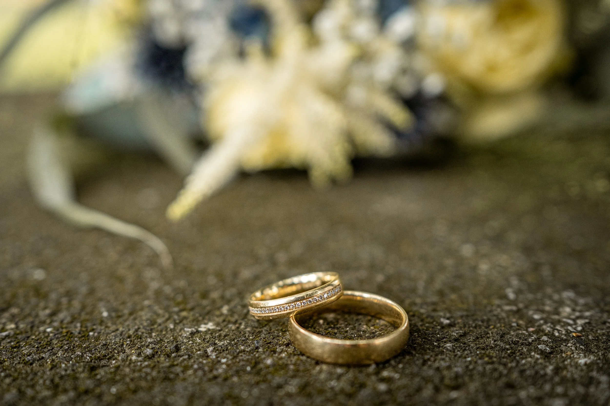 Zwei goldene Eheringe auf dunklem Sand, im Hintergrund unscharf ein verrosteter Gegenstand.