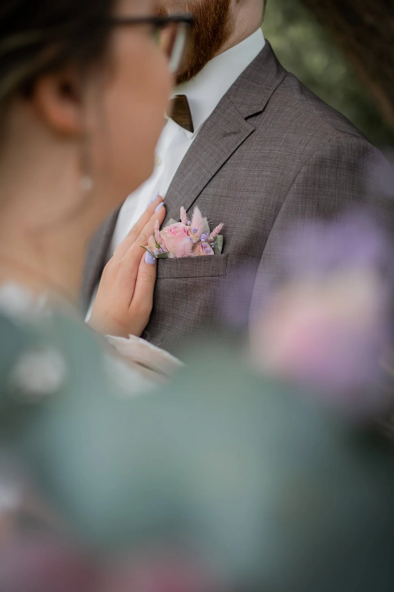 Nahaufnahme eines Mannes in Anzug mit Blumen im Revers bei einer Hochzeit, Nahaufnahme von Schulter und Brust. Die Frau ist leicht unscharf im Vordergrund zu sehen.