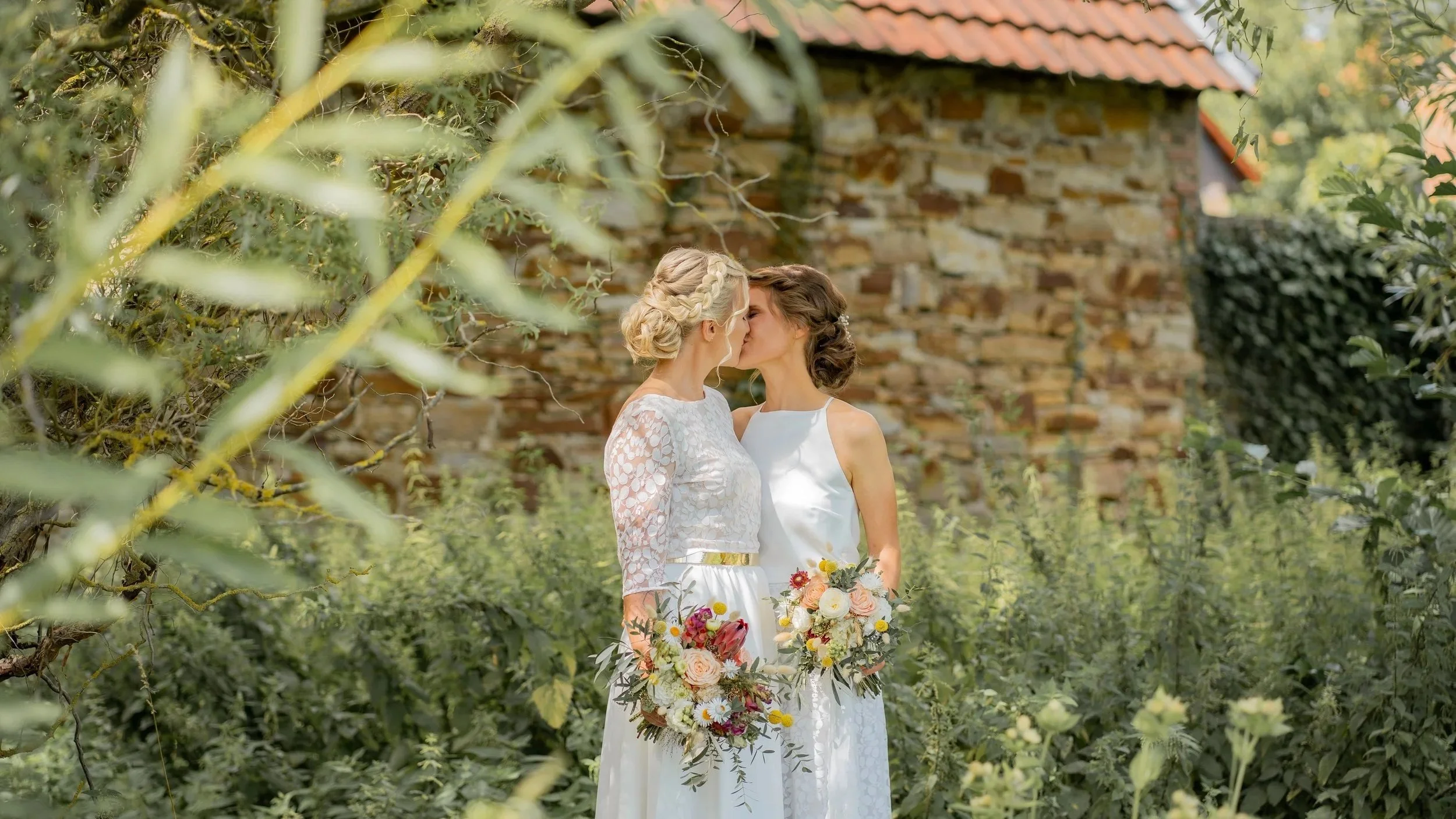 Zwei Frauen bei einer Hochzeit im Garten, küssen sich, im Hintergrund eine alte Steinmauer
