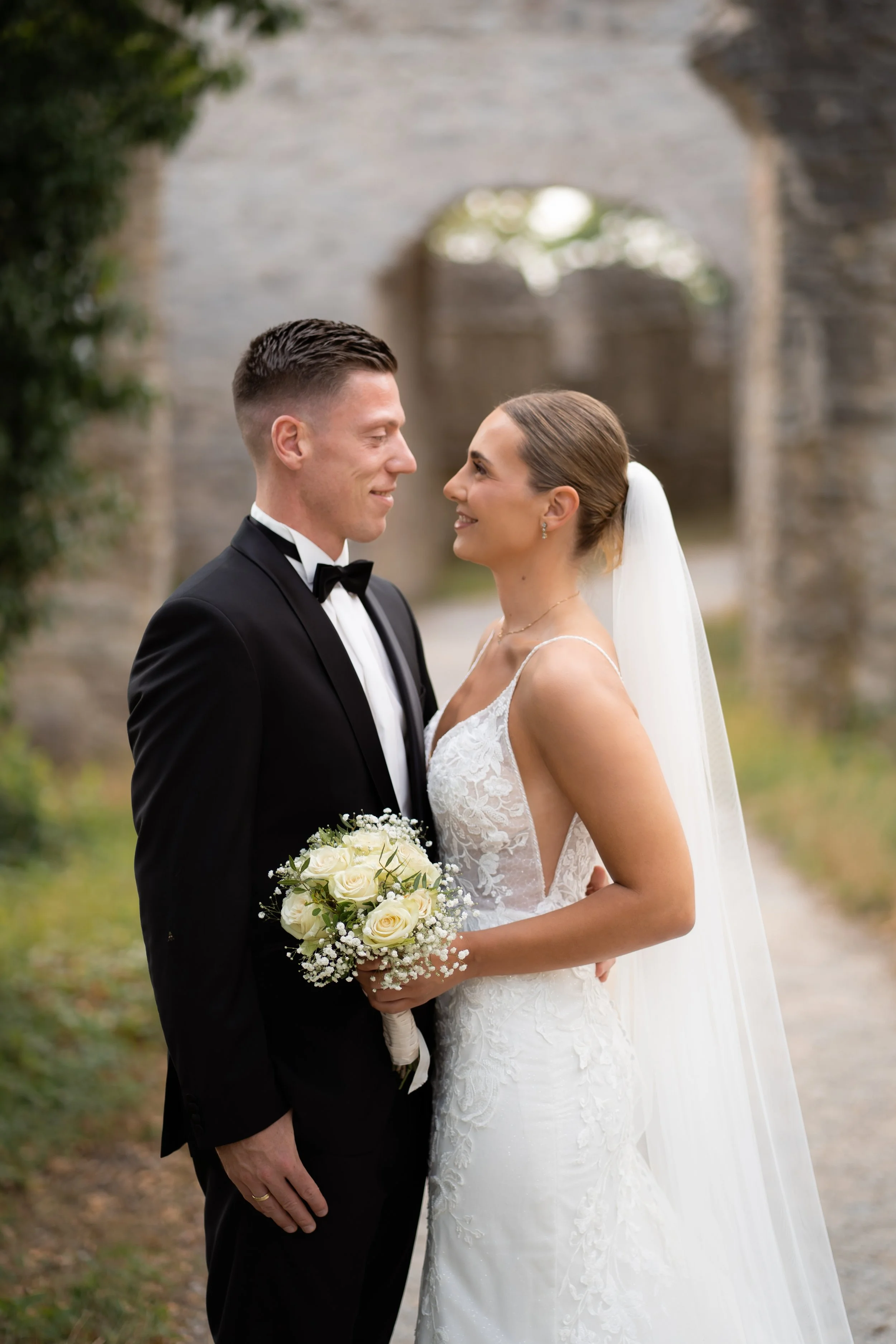 Ein Brautpaar steht sich bei einer Hochzeit im Freien gegenüber. Der Bräutigam trägt einen schwarzen Anzug mit Fliege, die Braut trägt ein weißes Brautkleid mit Spitzen und einen Schleier. Sie hält einen Blumenstrauß mit weißen Rosen. Im Hintergrund 