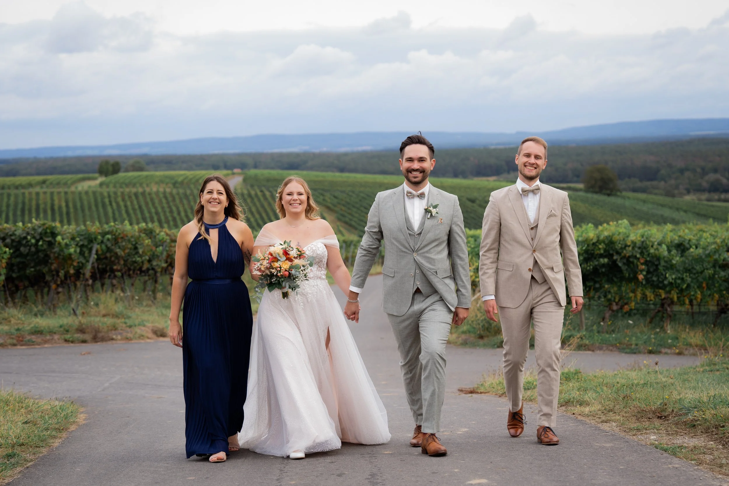Eine Gruppe von fünf Menschen bei einer Hochzeit, darunter das Brautpaar in der Mitte, umgeben von Freunden oder Familienmitgliedern, die im Freien auf einer Landstraße zwischen Weinbergen unterwegs sind.