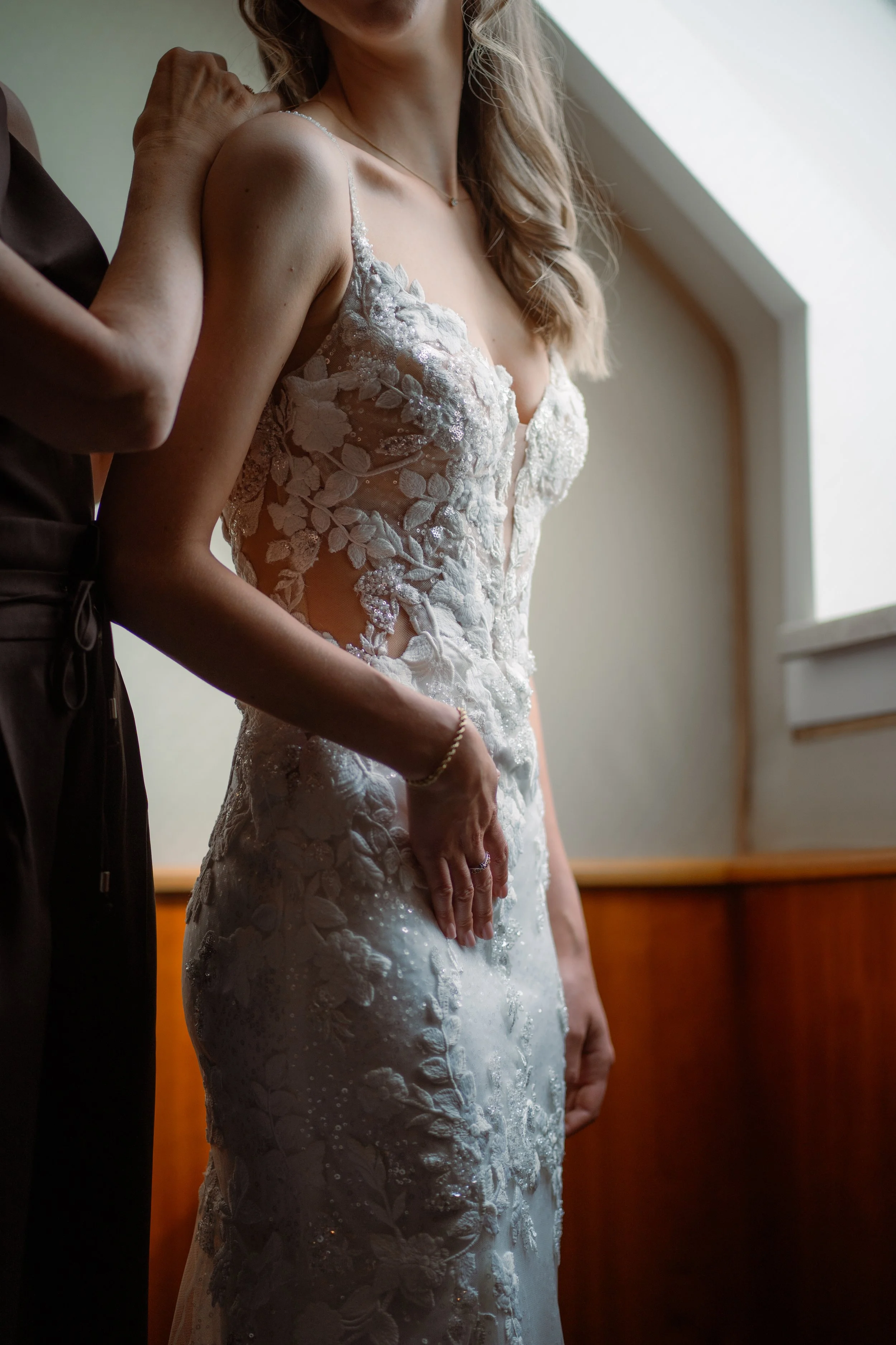 Nahaufnahme einer Frau im Brautkleid, die von einer anderen Person unterstützt wird, während sie sich für eine Hochzeit vorbereitet.