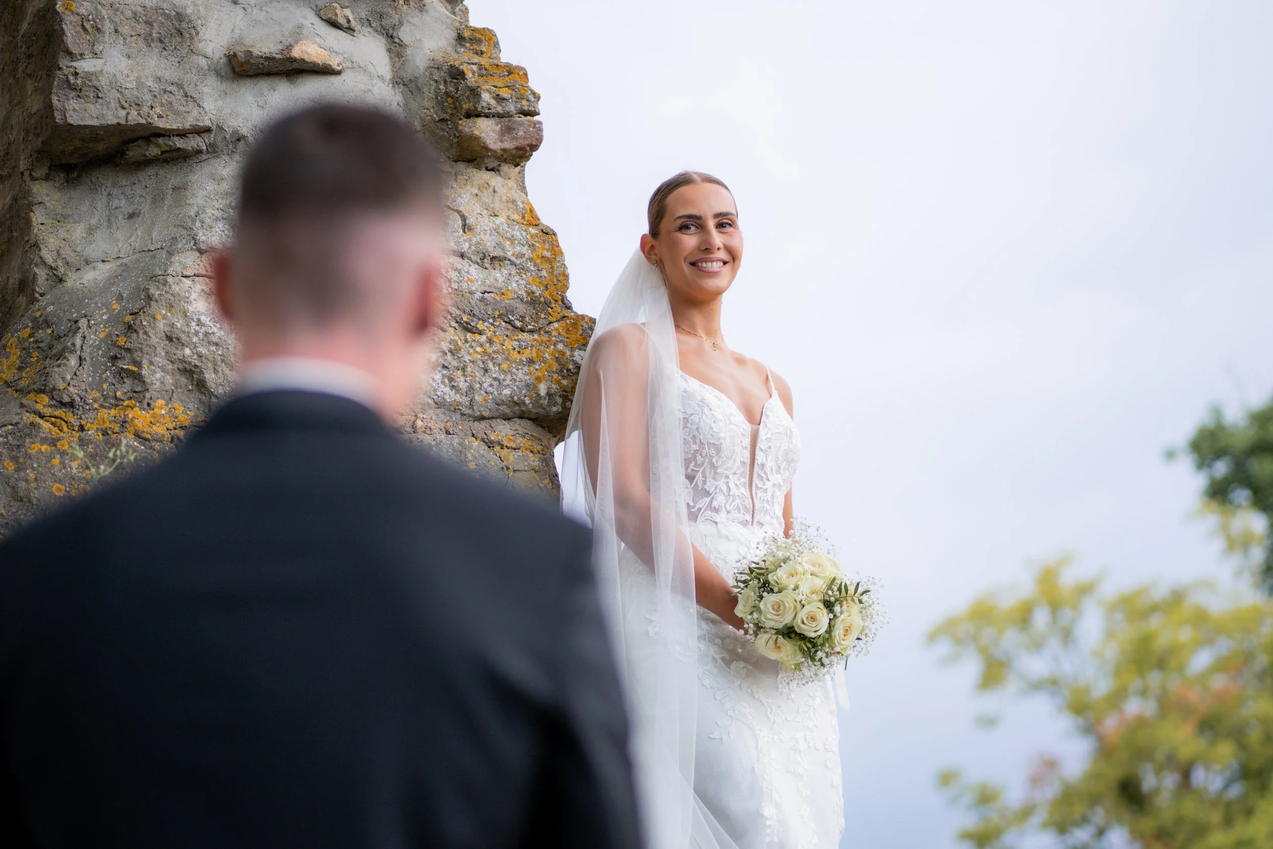 Braut in weißem Kleid hält Blumenstrauß bei Hochzeit im freien, Blick auf den Bräutigam im Vordergrund, hoher Himmel, Herbstbäume im Hintergrund.