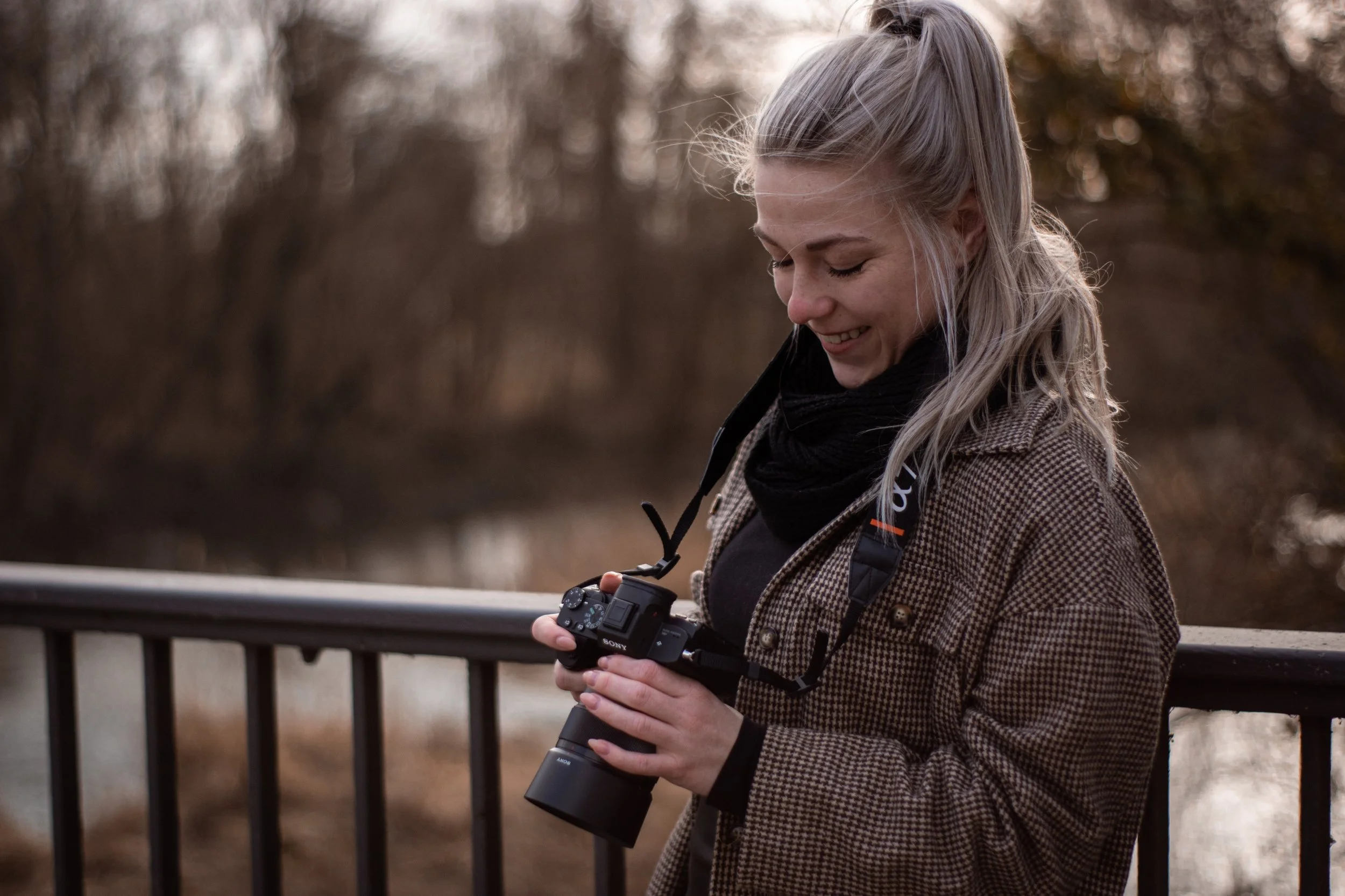 Eine lachende junge Frau mit blonden Haaren in einem Zopf, die eine Kamera in den Händen hält, im Freien auf einer Brücke, im Hintergrund Herbstlaub und Bäume.