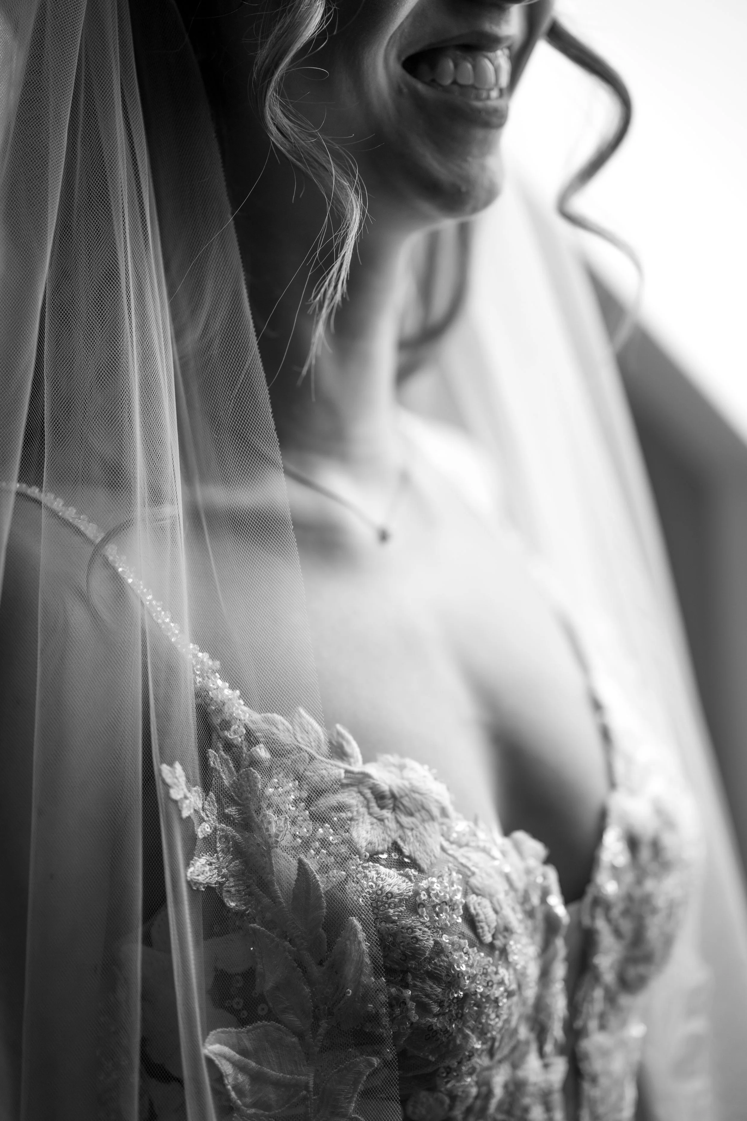 Schwarz-weiß-Foto eines lachenden Mädchens in einem weißen Kleid mit handarbeitverzierter Spitze, getragen zu einem besonderen Anlass. Im Vordergrund ist ein Schleier zu sehen.