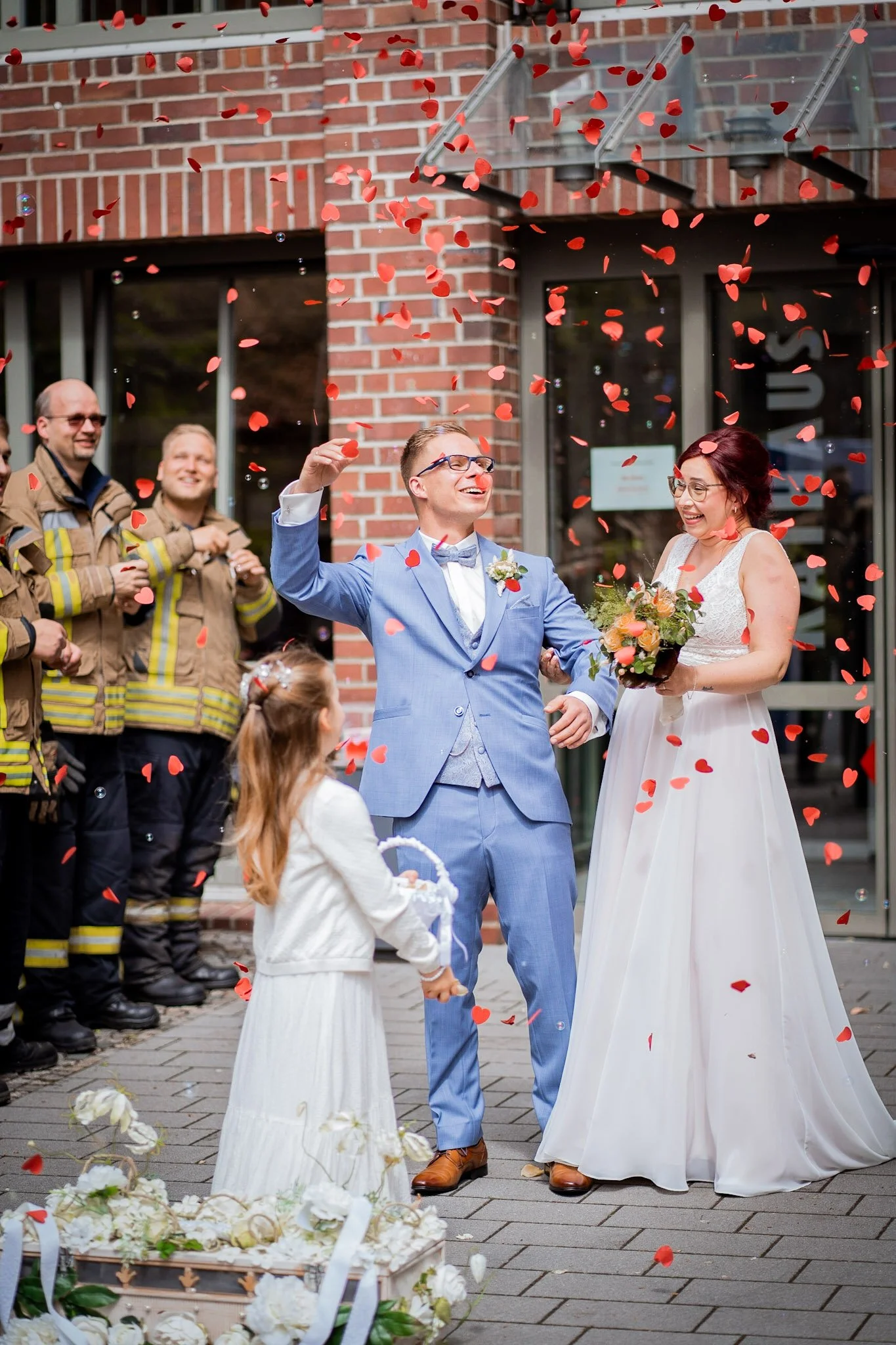 Ein Hochzeitsfest mit lachenden Menschen, die Blumen und Herzkonfetti werfen, vor einem Backstein-Gebäude.