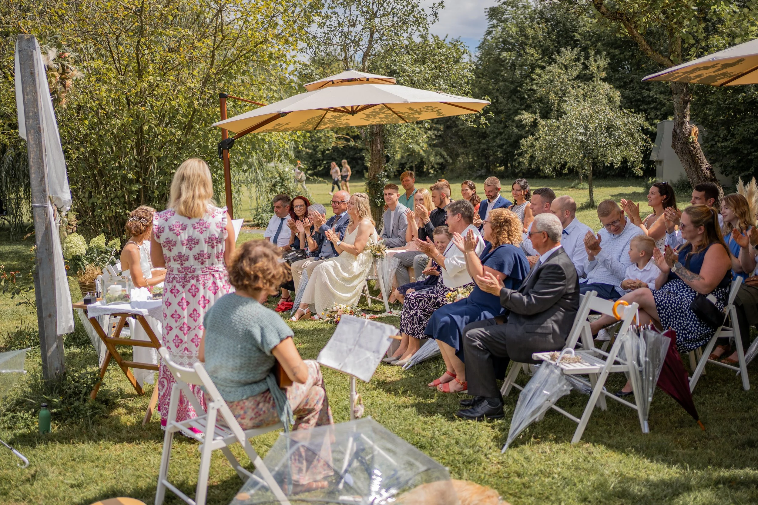 Eine Gruppe von Menschen sitzt bei einer Hochzeit im Freien unter Sonnenschirmen, umgeben von Bäumen und grünem Gras. Es findet eine Zeremonie statt, bei der eine Frau eine Rede hält, während die Gäste applaudieren und lächeln.