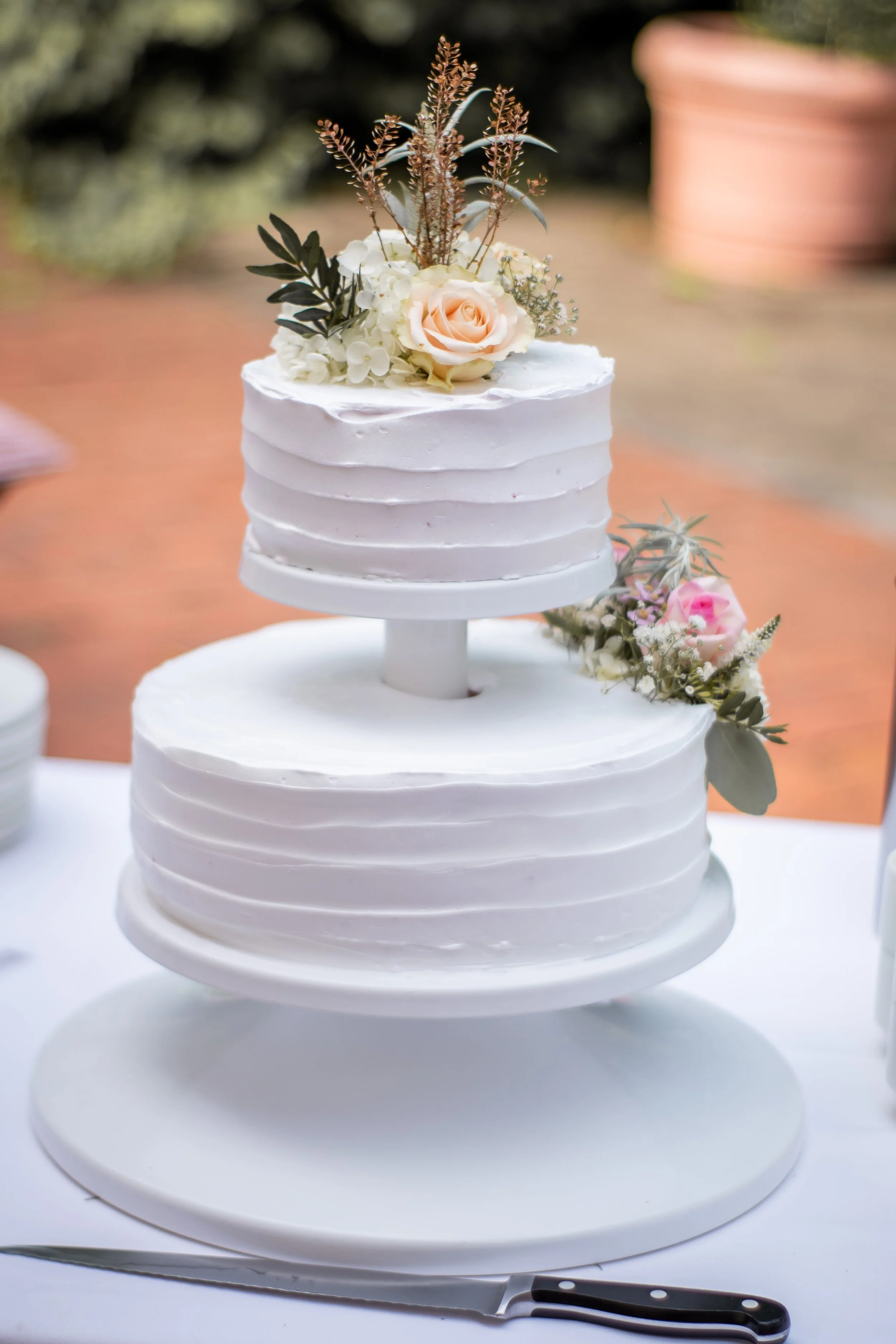 Ein mehrstöckiger weißer Hochzeitstorte mit Blumen auf der oberen Etage, auf einem weißen Teller. Ein Messer liegt daneben.