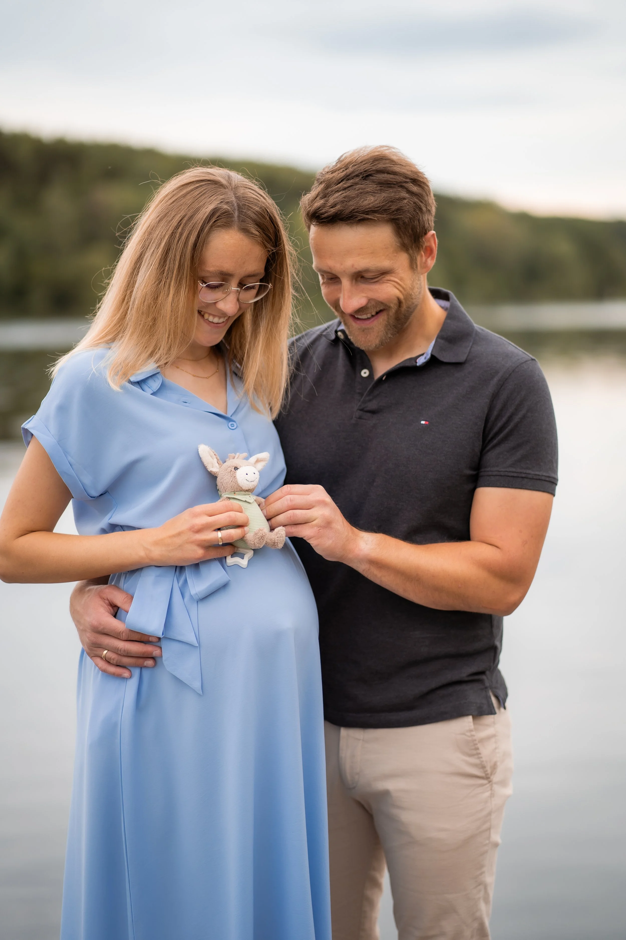 Ein schwangeres Paar steht am Fluss, der Mann hält die Hand seiner Partnerin und beide schauen lachend auf ein kleines Stofftier in den Händen der Frau.