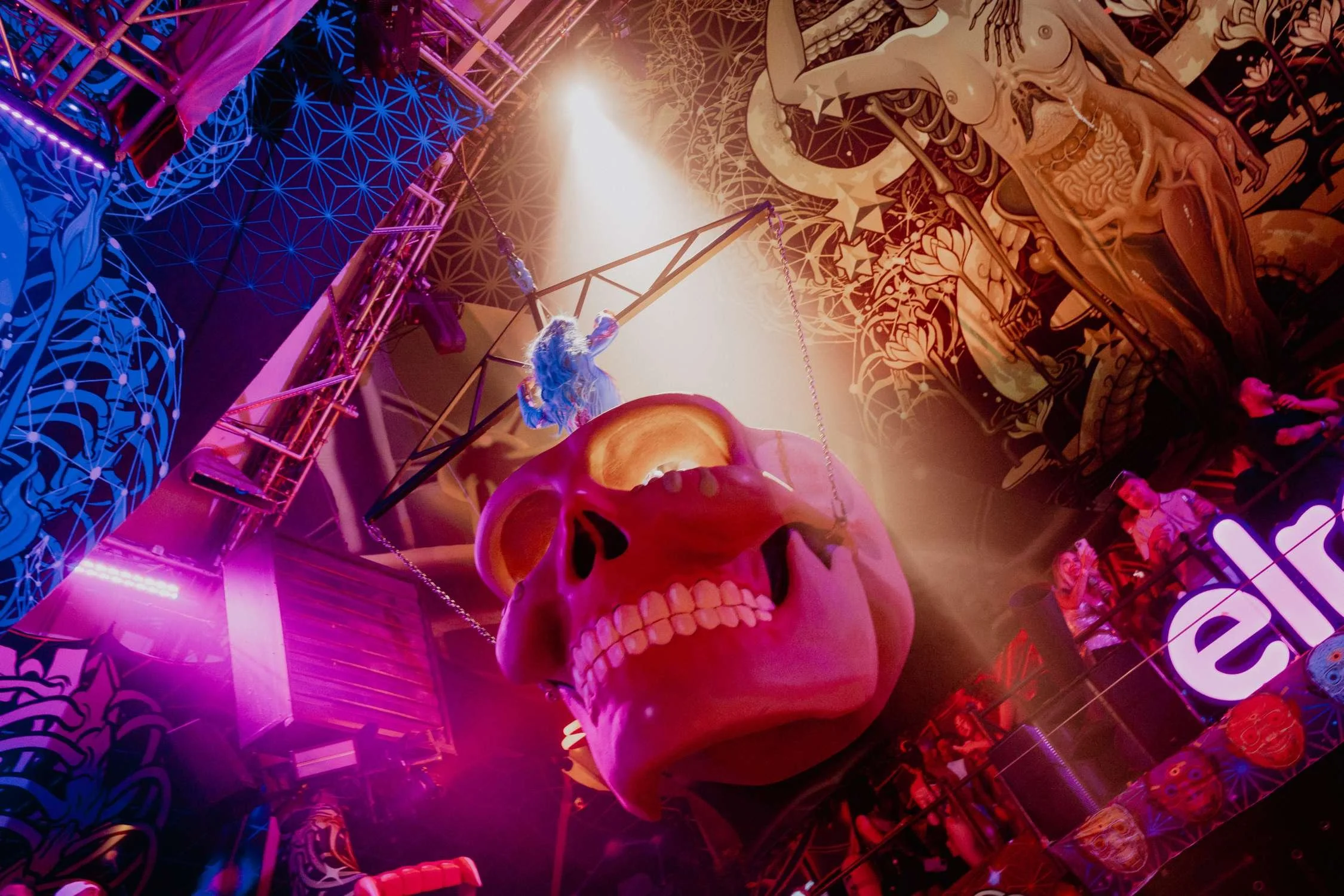 Aerial performer above a giant fluorescent skull set piece inside an immersive club environment with UV lighting.