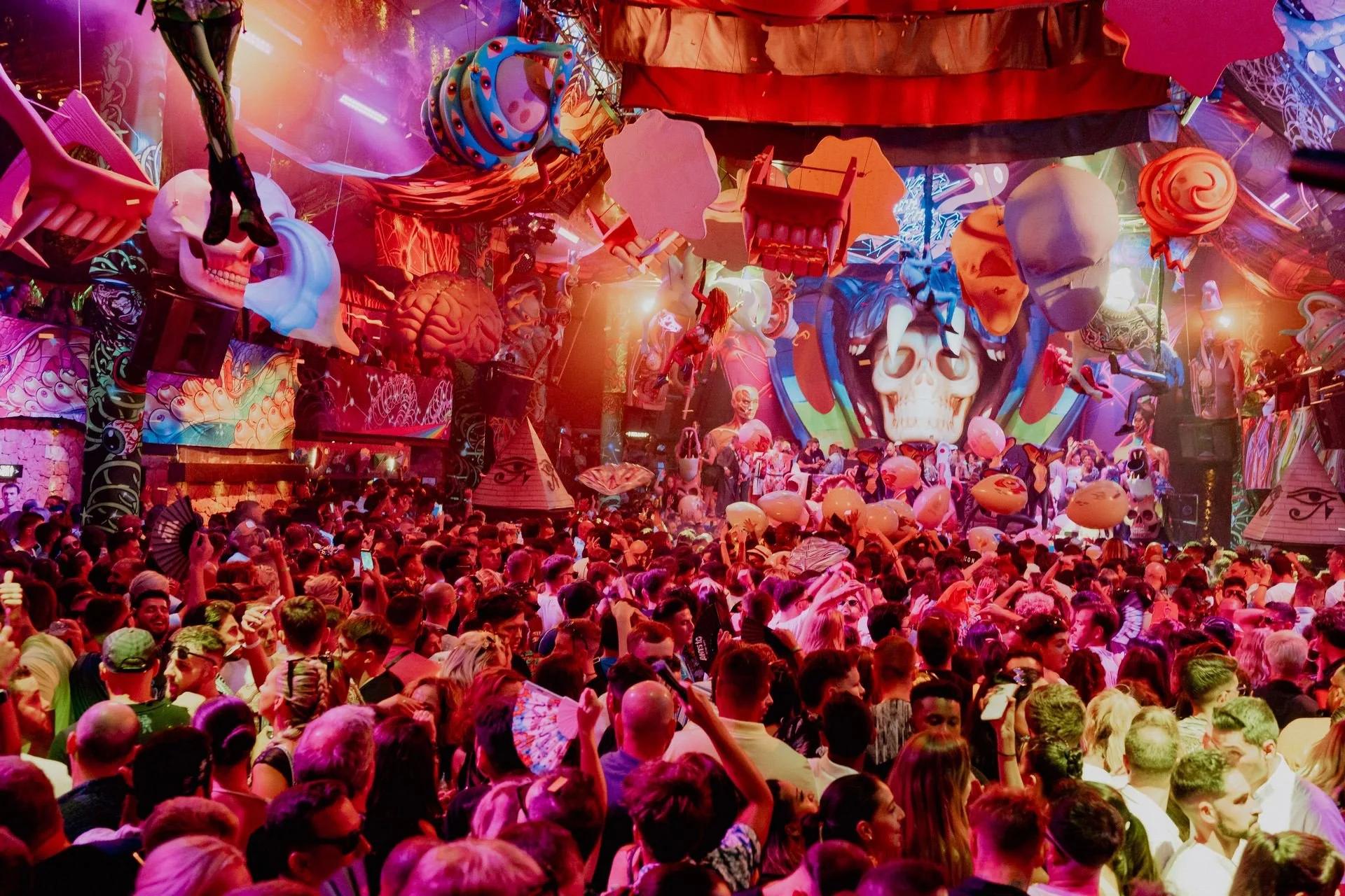 Packed dance floor facing an immersive stage with hanging inflatables, skull centerpiece, and UV lighting effects.