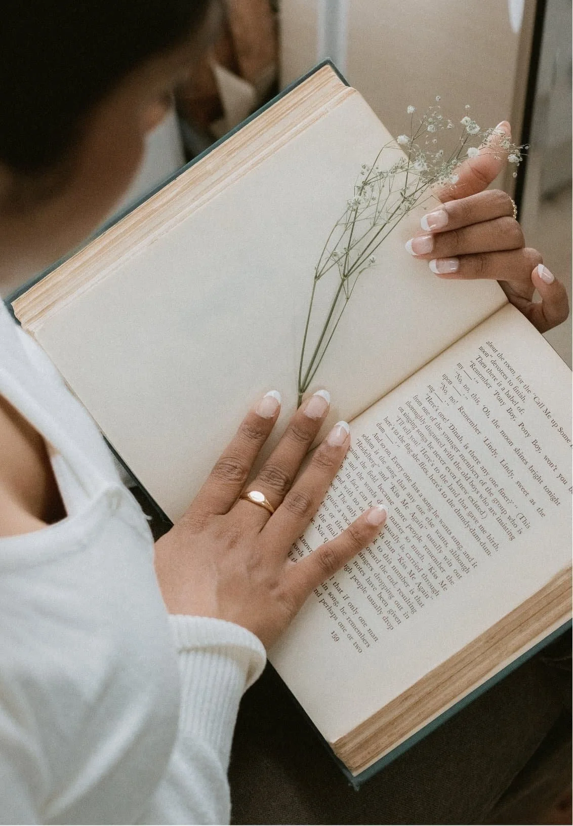 A woman with neatly manicured nails and a gold ring holds an open book, using a sprig of small white flowers as a bookmark. The book on her lap hints at quiet reflection, perhaps exploring concepts like IFS therapy, while she wears a white top.
