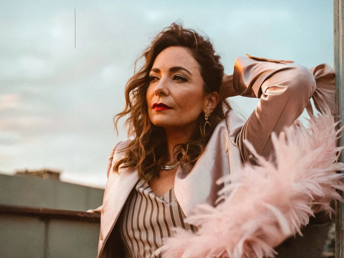 Elegant woman with wavy brown hair in a blazer, gazing thoughtfully outdoors. Warm lighting and stylish accessories enhance her look.