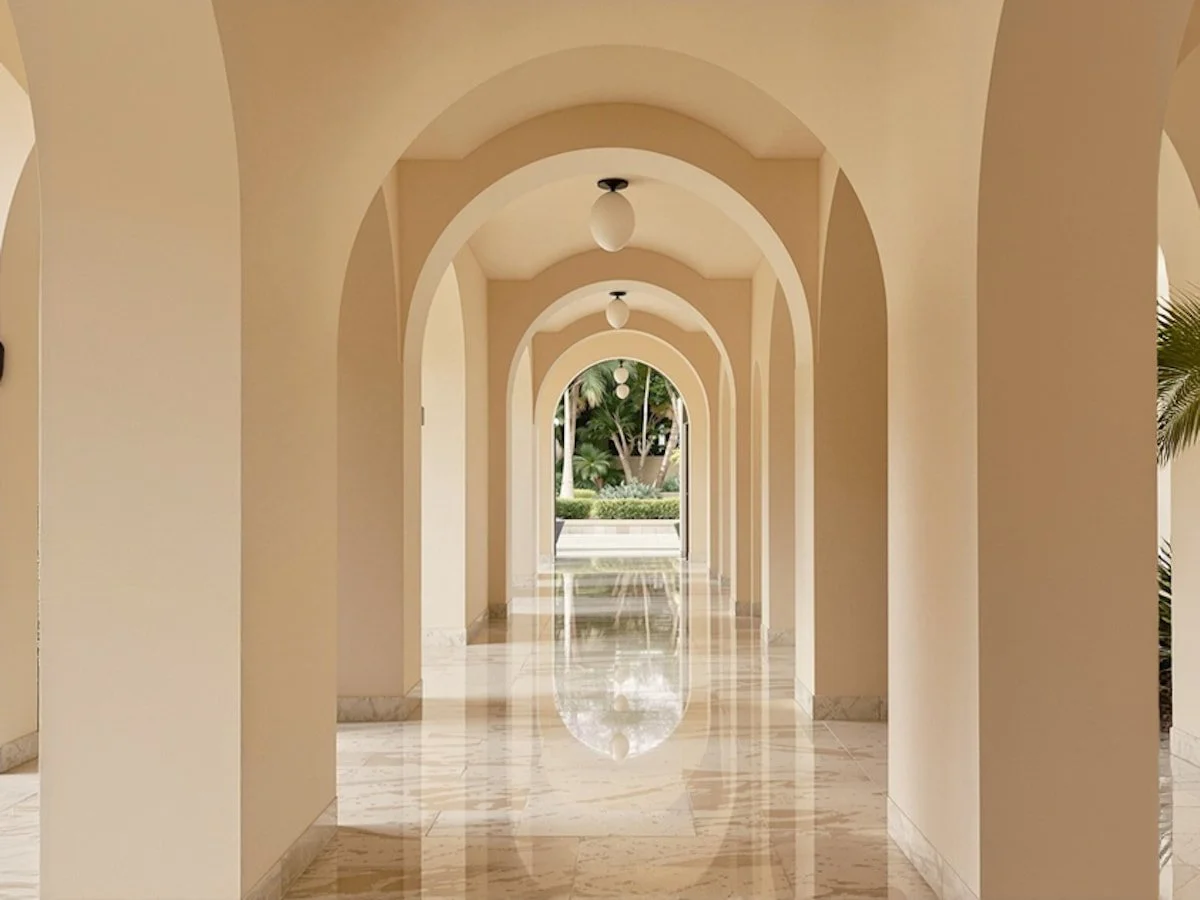 Elegant arched hallway with polished marble floors, warm lighting, and lush greenery visible at the end. A serene architectural design.