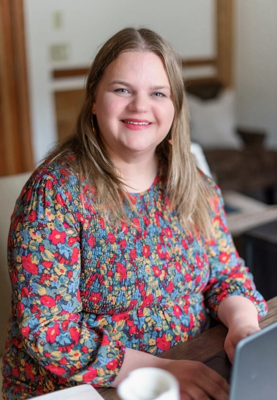 Morgen Villegas, with long, light brown hair and a colorful floral blouse, smiles while sitting at a table and working on a laptop. The softly blurred background creates a warm, inviting atmosphere.