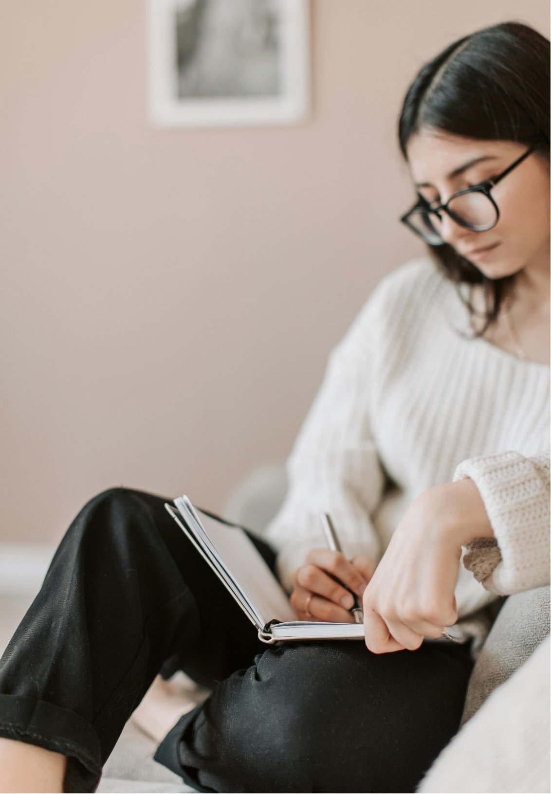 A woman with long dark hair and glasses, wearing a white sweater and black pants, sits indoors and writes in a notebook while looking down, focused on her IFS therapy work.