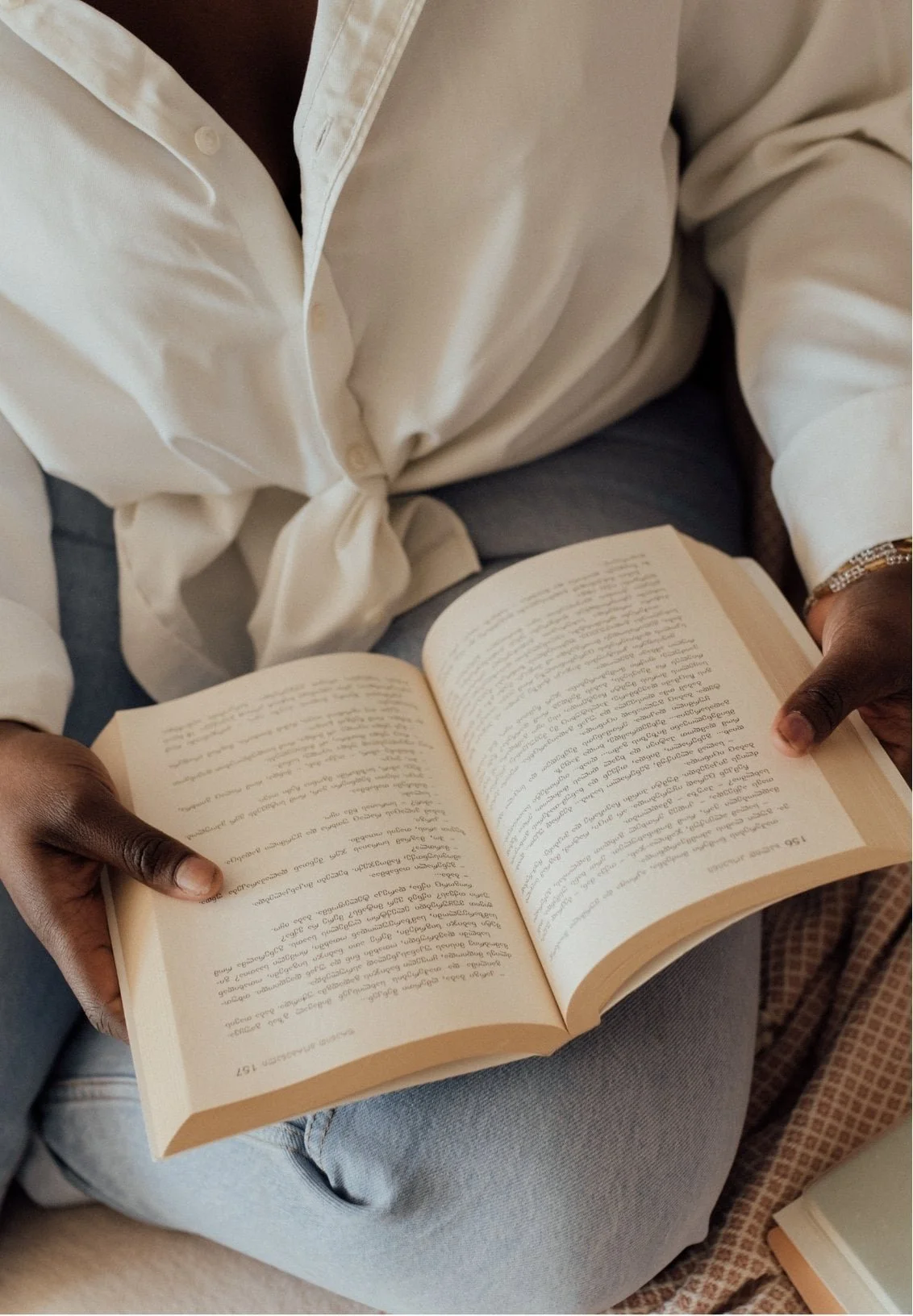 A woman wearing a white shirt and light blue jeans is sitting cross-legged, reading an open book—possibly about therapy for anxiety—holding it with both hands. Only her torso and hands are visible.