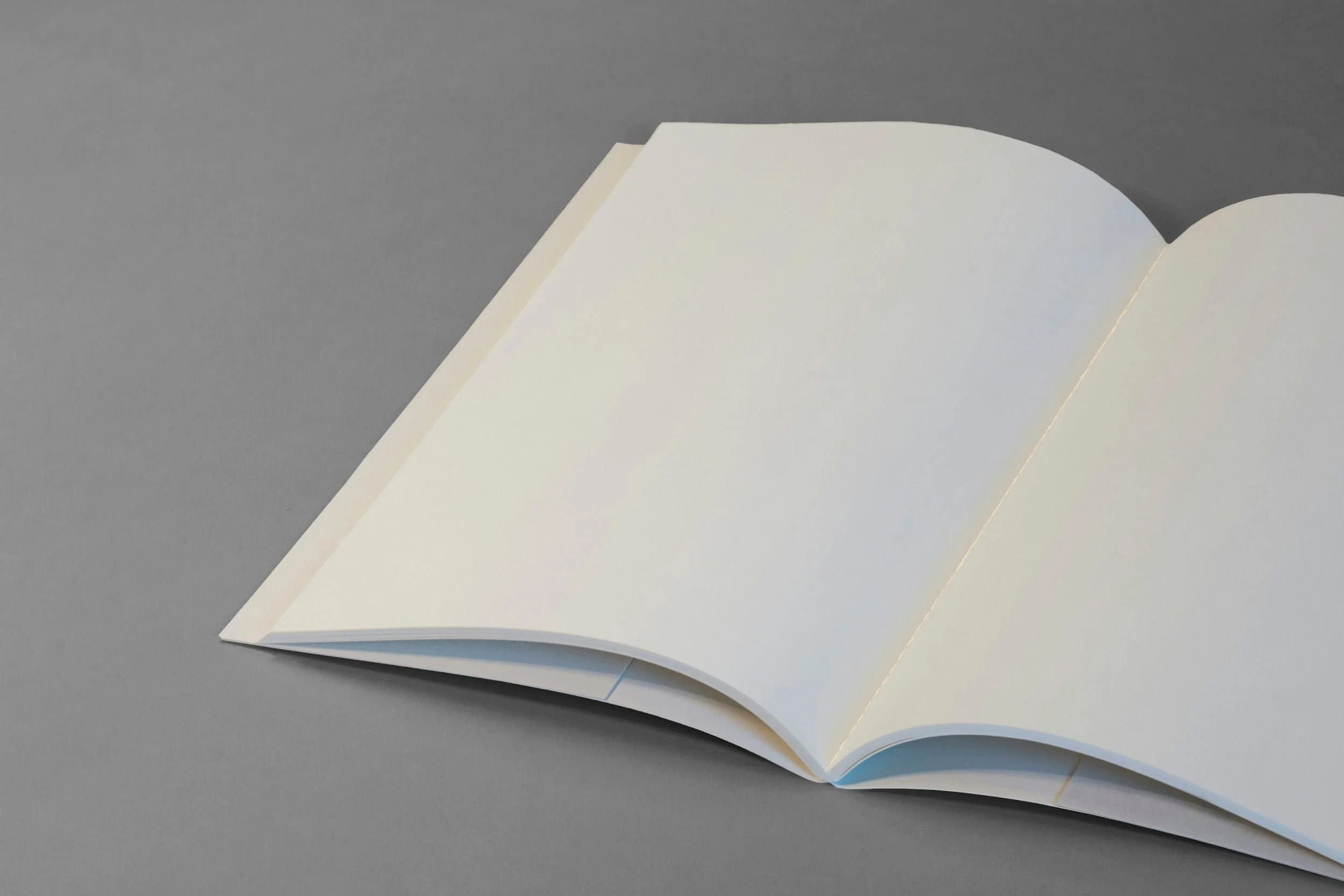 An open blank book with white pages lies on a flat, gray surface. The book is angled slightly, showing both pages and part of the books spine.