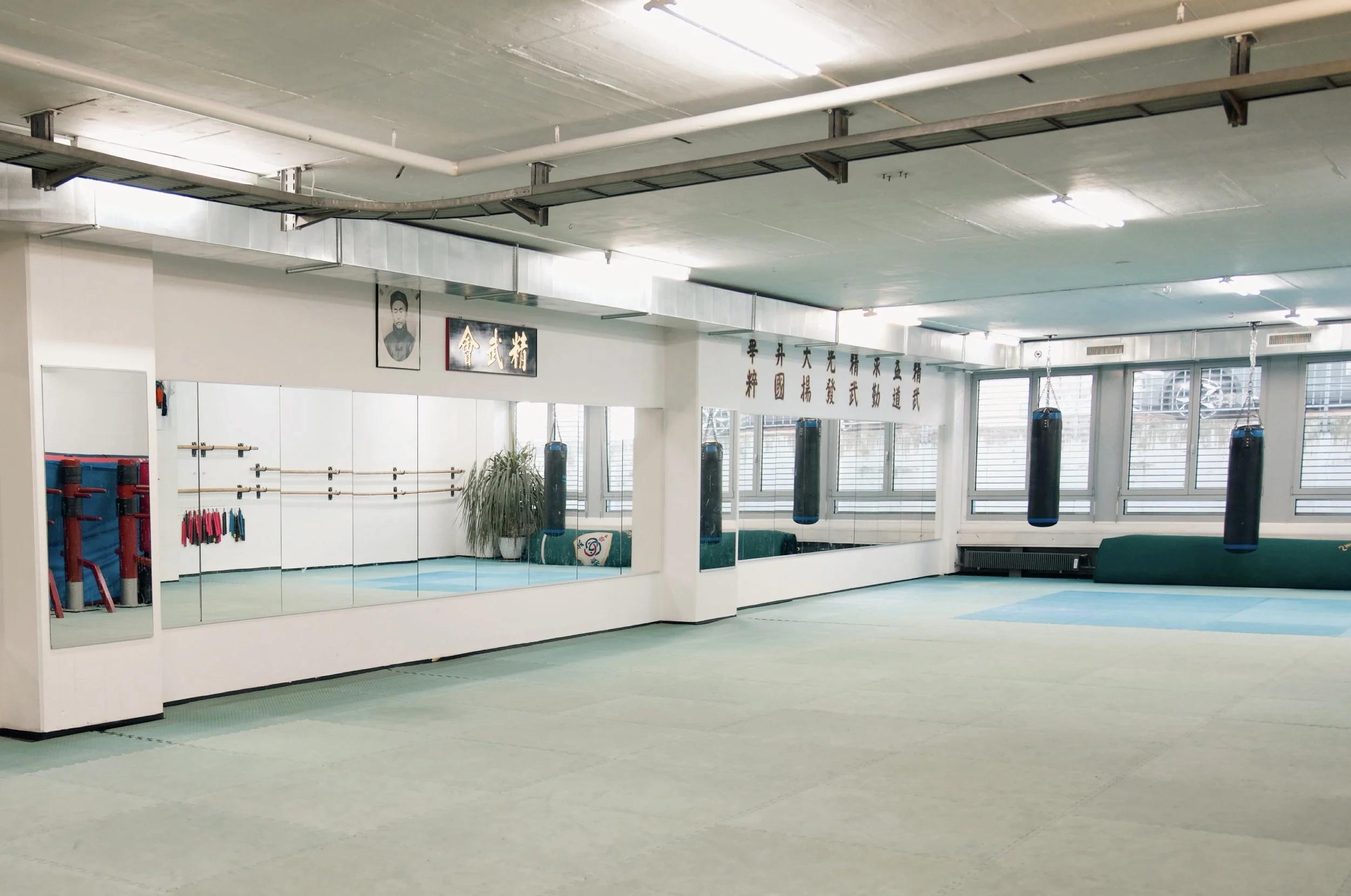 Leeres Kampfsporttrainingraum mit Spiegelwand, Boxsäcken, Sonnenlicht durch große Fenster, Wand mit chinesischen Schriftzeichen.