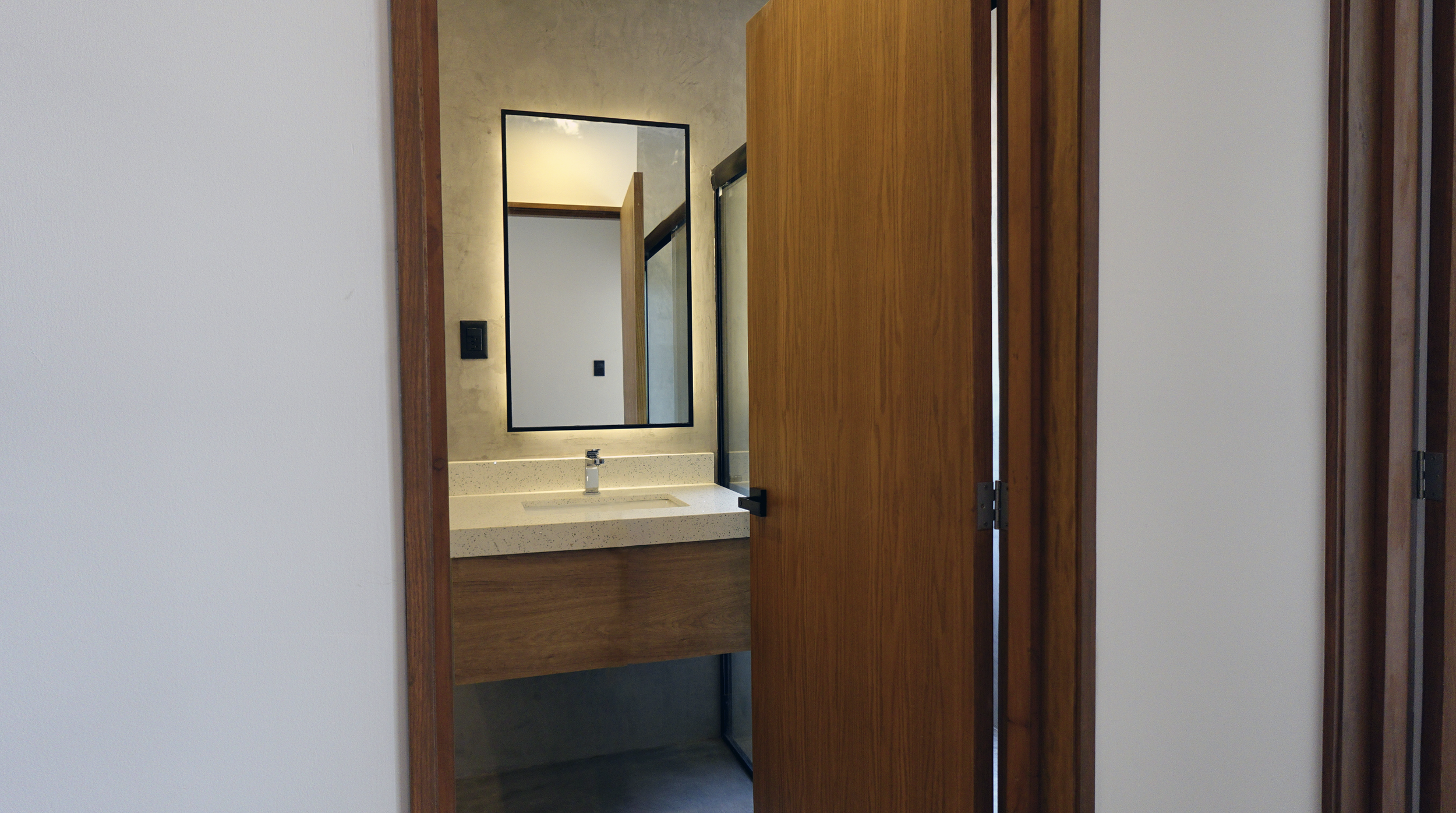 Vista de un baño con espejo, lavabo de granito y paredes de cemento, visto a través de una puerta de madera.