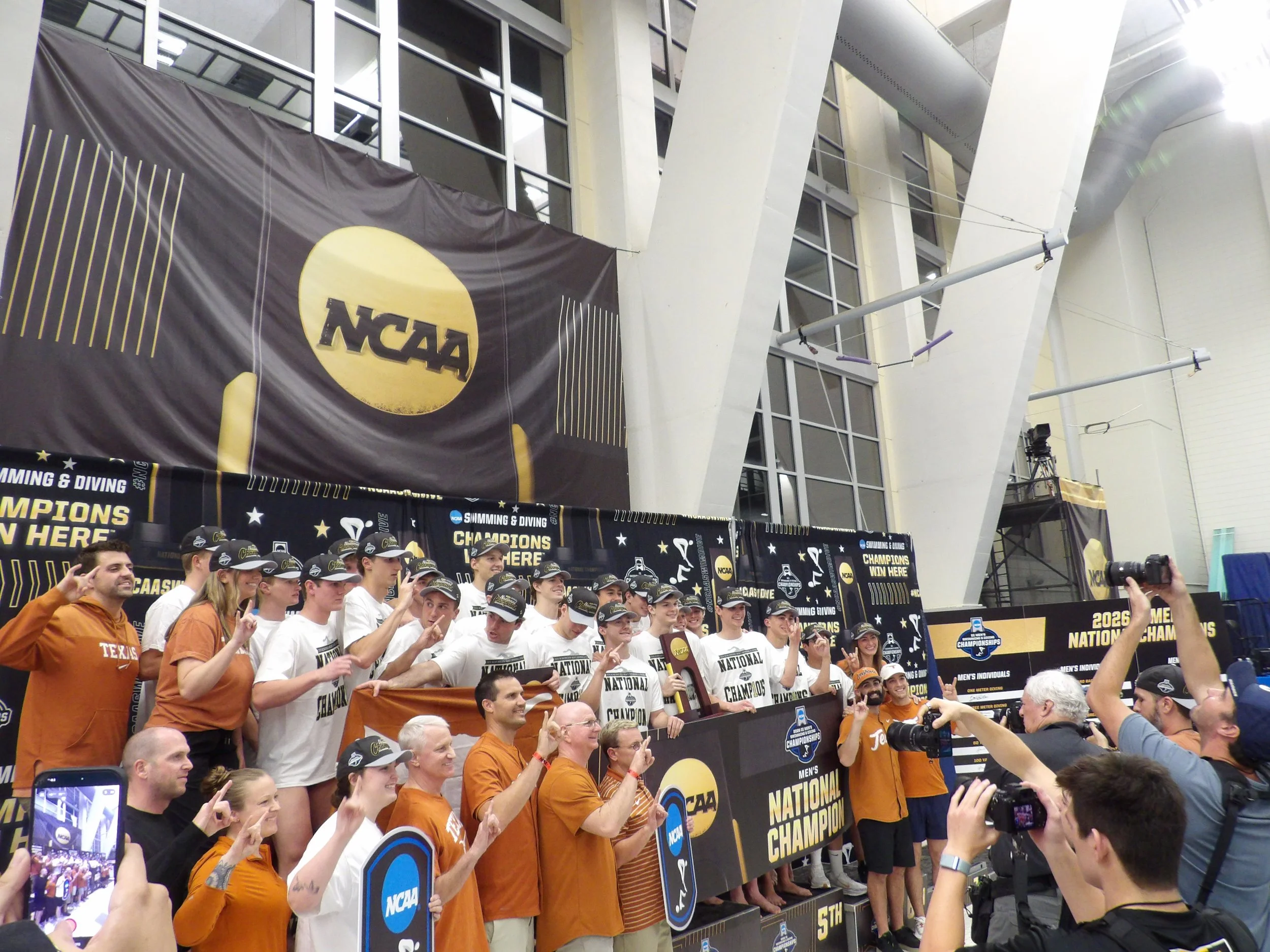 A group of Texas swimmers and coach Bob Bowman celebrating at the NCAA podium with NCAA banners, indicating a national championship win in swimming and diving, in an indoor stadium with large windows and modern architecture.