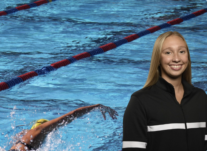 A young female swimmer in a pool with a swimming lane, smiling while wearing a black jacket with white stripes.