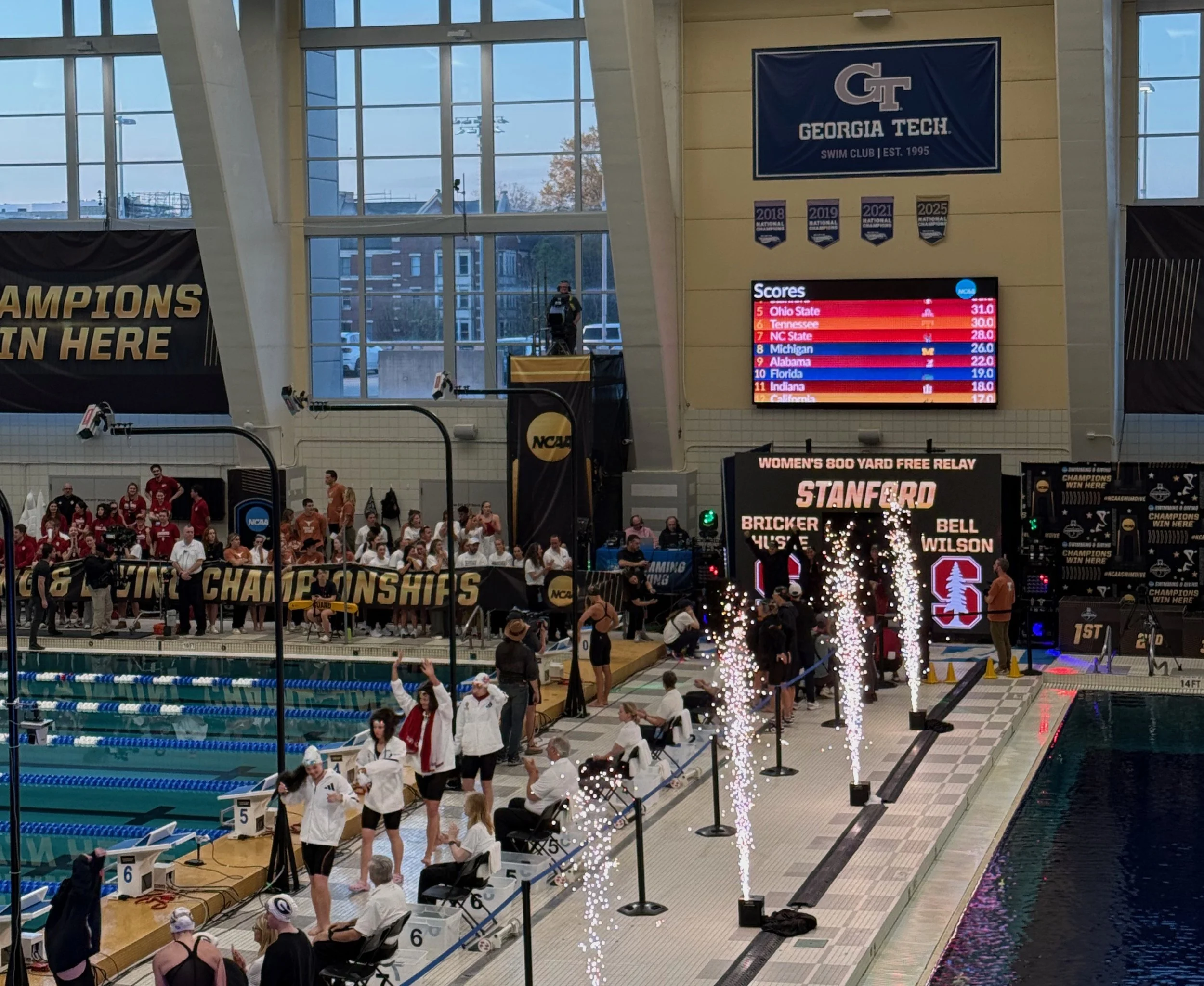 Indoor swimming pool area with swimmers, spectators, and officials. Large digital scoreboard displays scores for women's 800 yard freestyle relay with teams and points. Georgia Tech banner, championship banners, and decorative fireworks are visible. Large windows with a cityscape outside.
