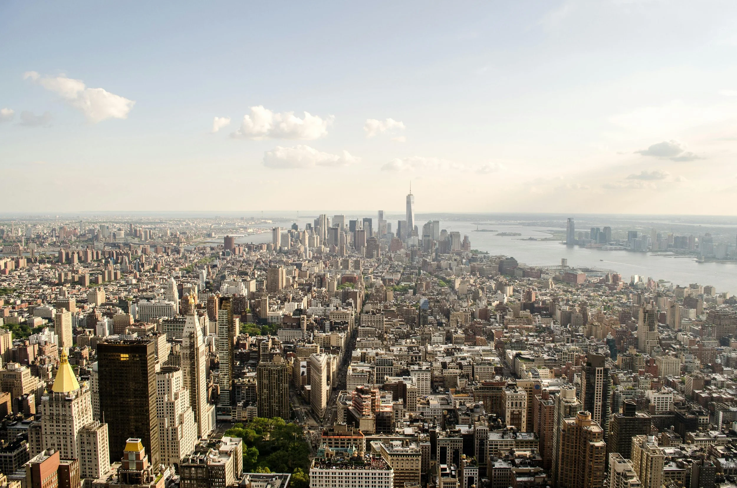 A city skyline featuring tall skyscrapers, a river on the right, and scattered clouds in the sky.